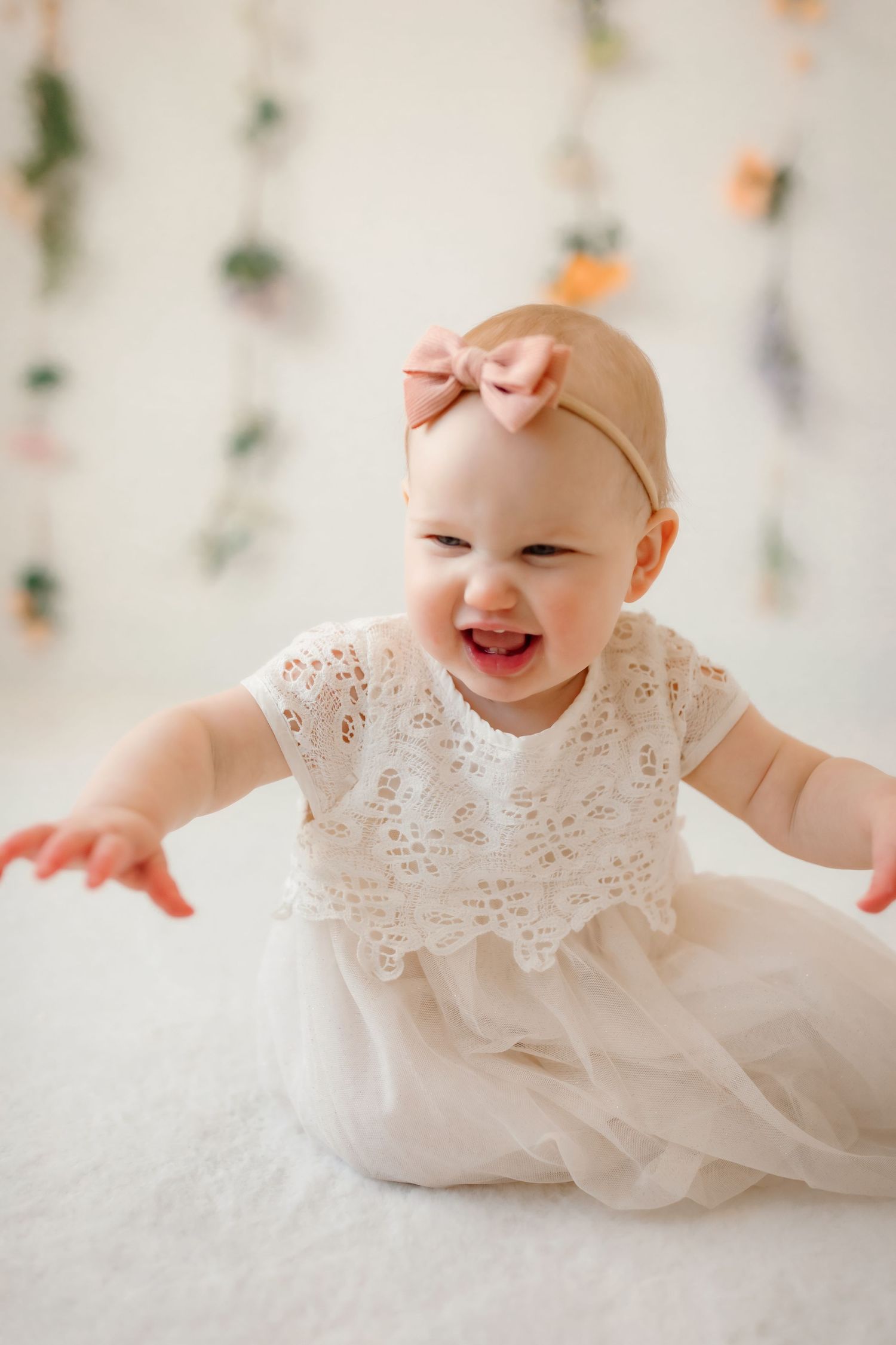 Baby in white lace dress with pink bow laughing against floral backdrop.