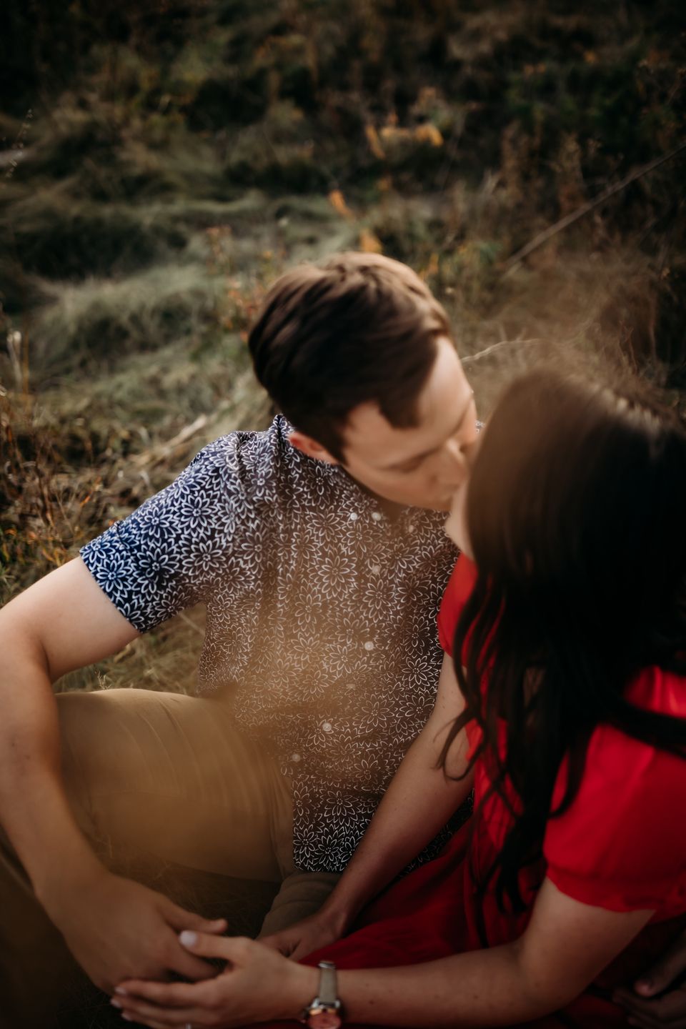 Tender embrace captured in sunlit field with couple in contrasting blue and red outfits.