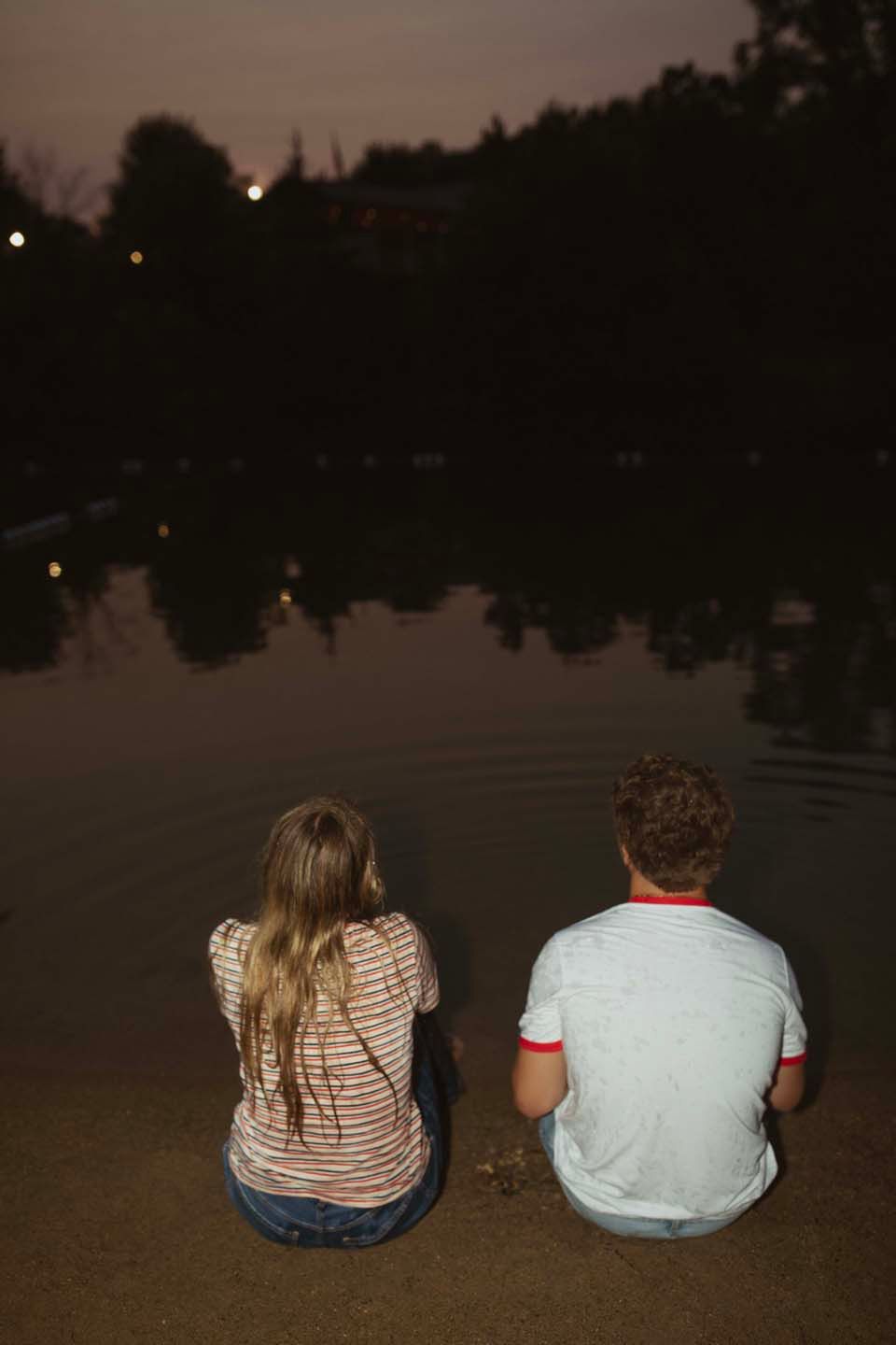 Two teenagers sit side by side at a pond's edge at night, gazing at the water.