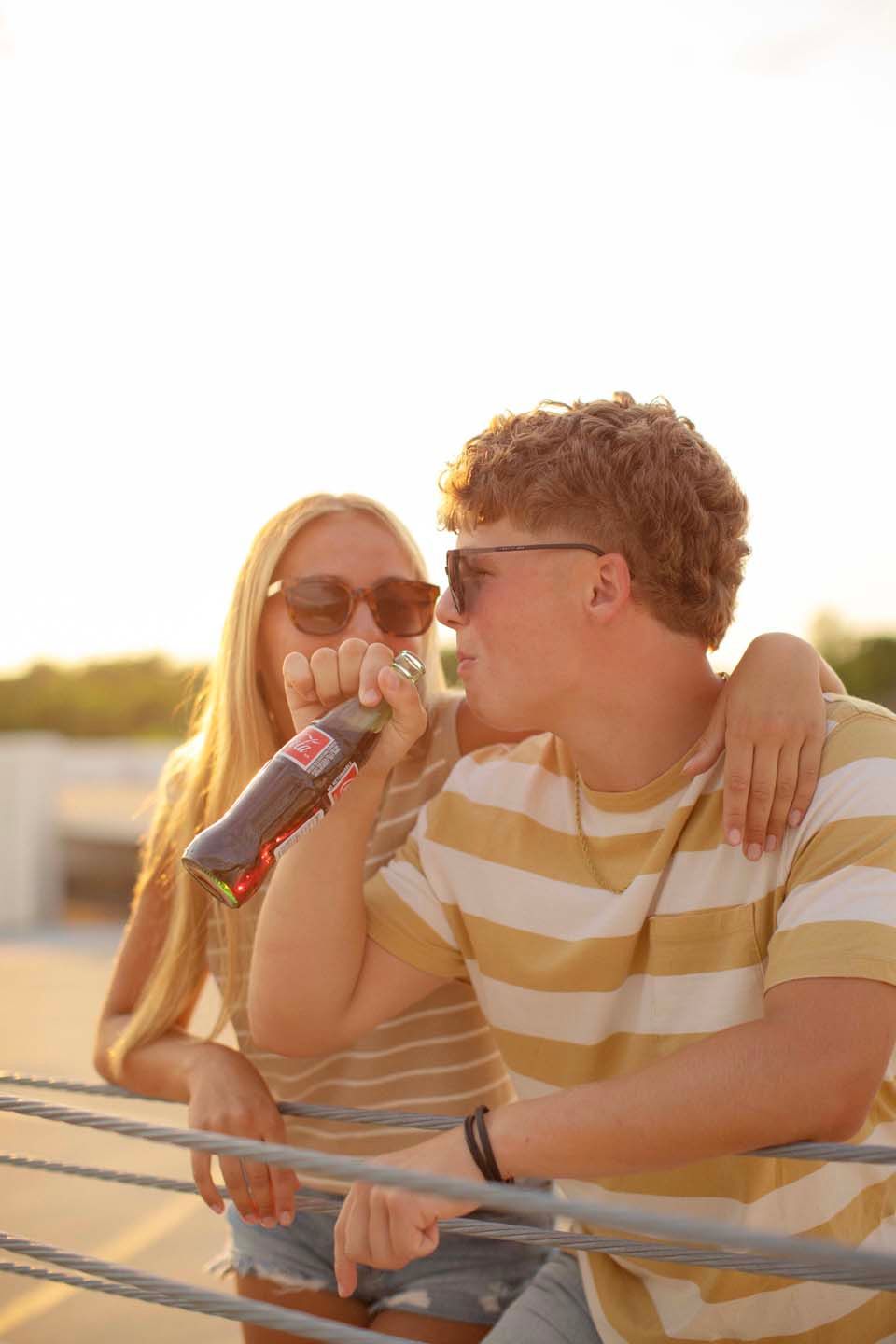 Young couple in sunglasses enjoying a soda drink together outdoors at sunset, leaning on a railing.