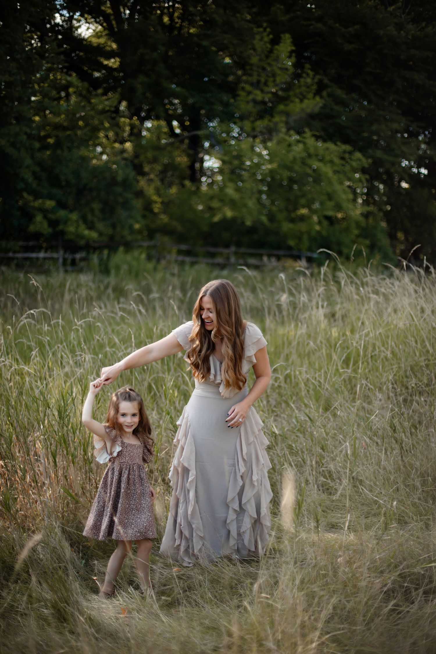 Mother and daughter dancing together in a golden meadow at sunset wearing flowing dresses among tall grasses.