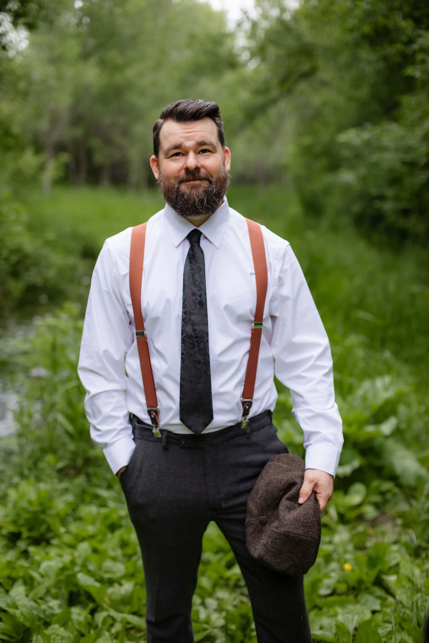 A groom in white shirt and brown suspenders poses casually in a lush green outdoor setting.