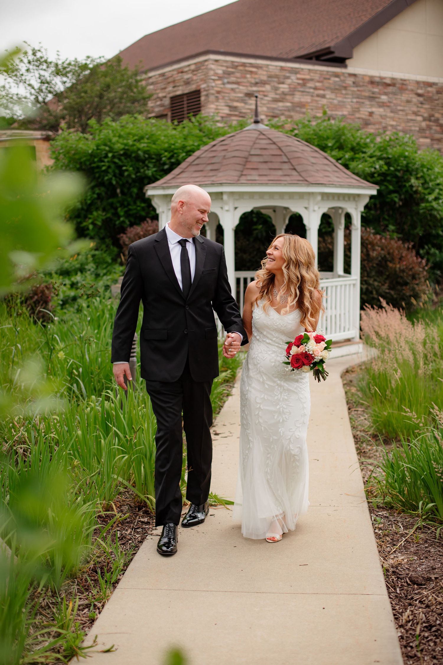 A couple walks along a garden path with a white gazebo and stone building in the background.