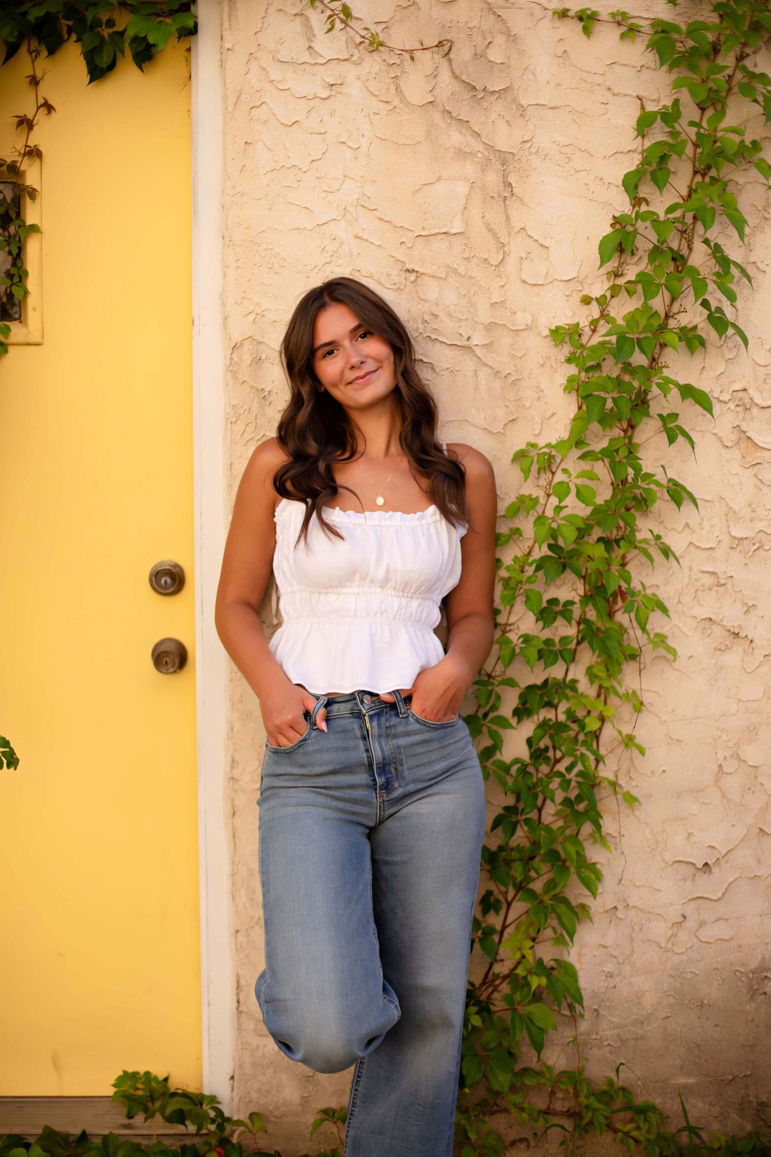 Young woman in white ruffled top and jeans posing by a yellow door with ivy-covered wall.