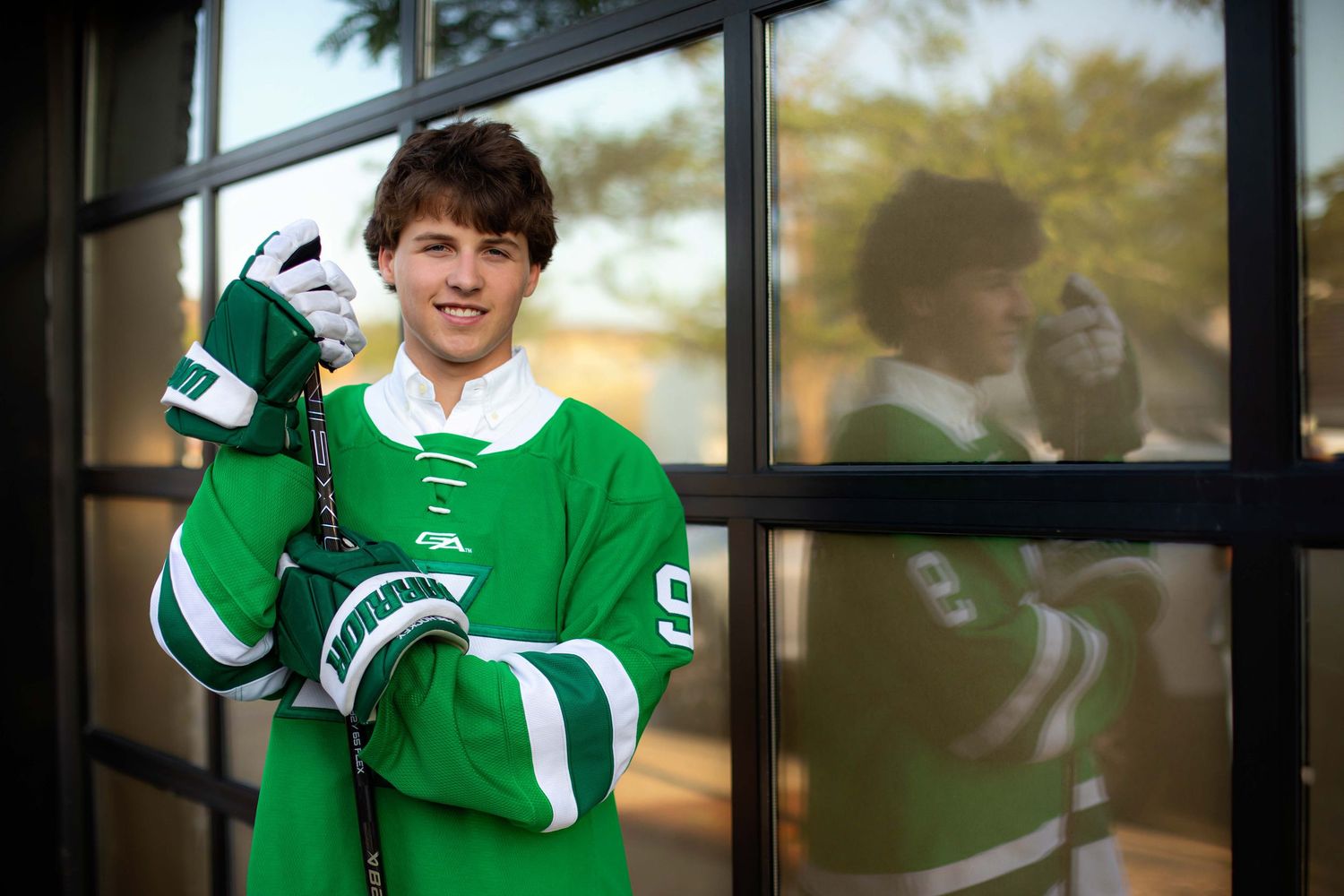 Young hockey player in green jersey holding stick, posing by glass doors with reflection visible.