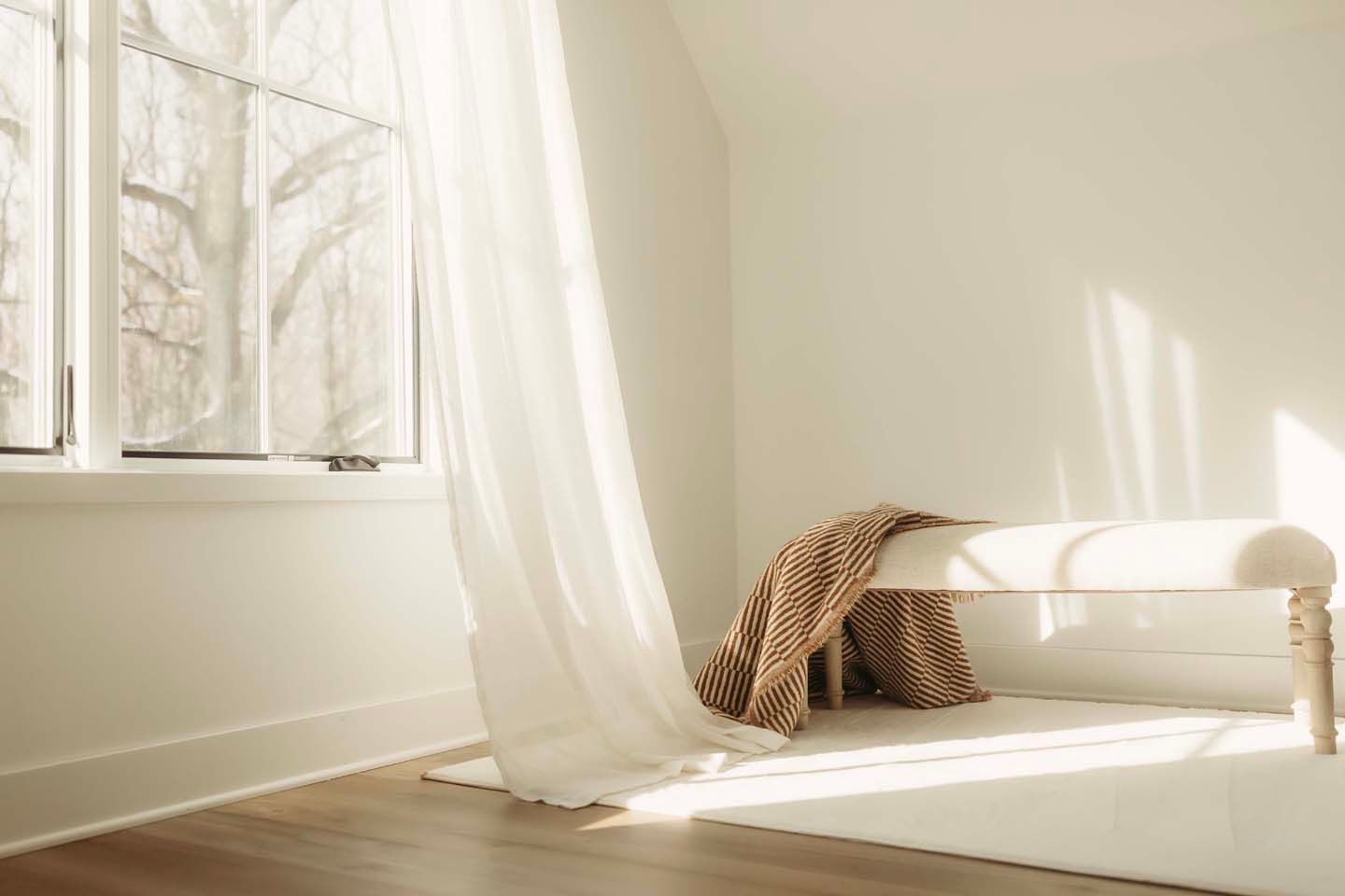 Bright sunlight streams through white sheer curtains into a minimalist room with hardwood floors.
