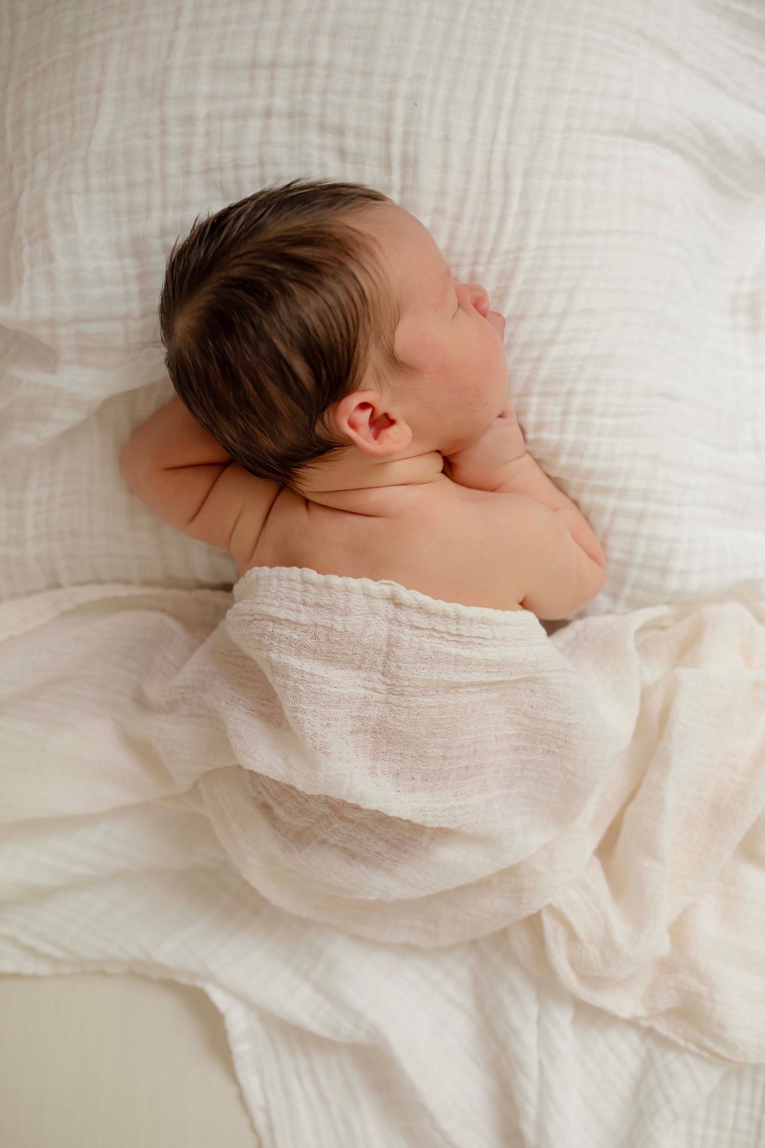 A peaceful newborn baby sleeping on a white pillow, wrapped in a soft muslin swaddle blanket.