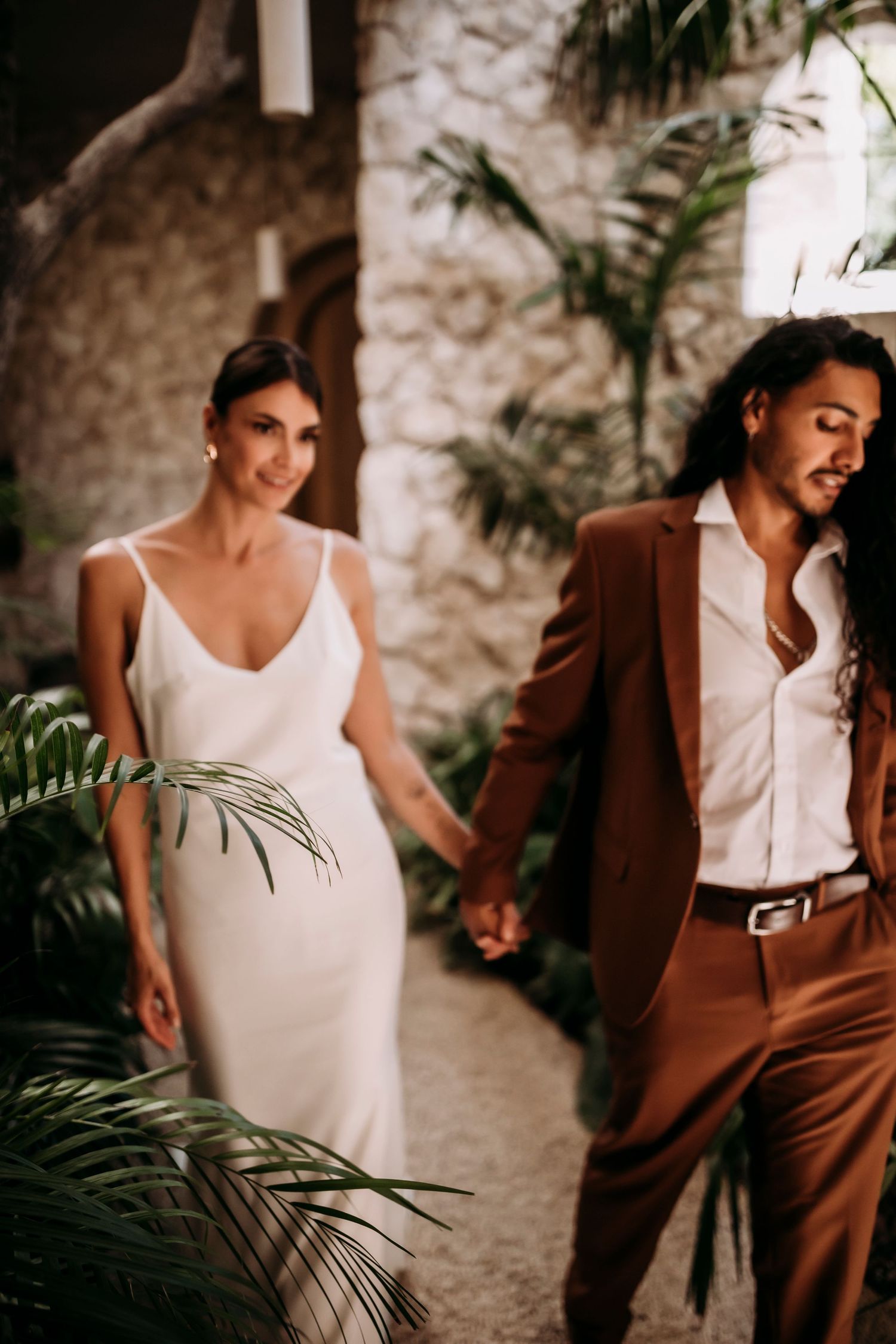 Elegant wedding couple stands among tropical palm fronds, bride in white slip dress and groom in brown suit holding hands.