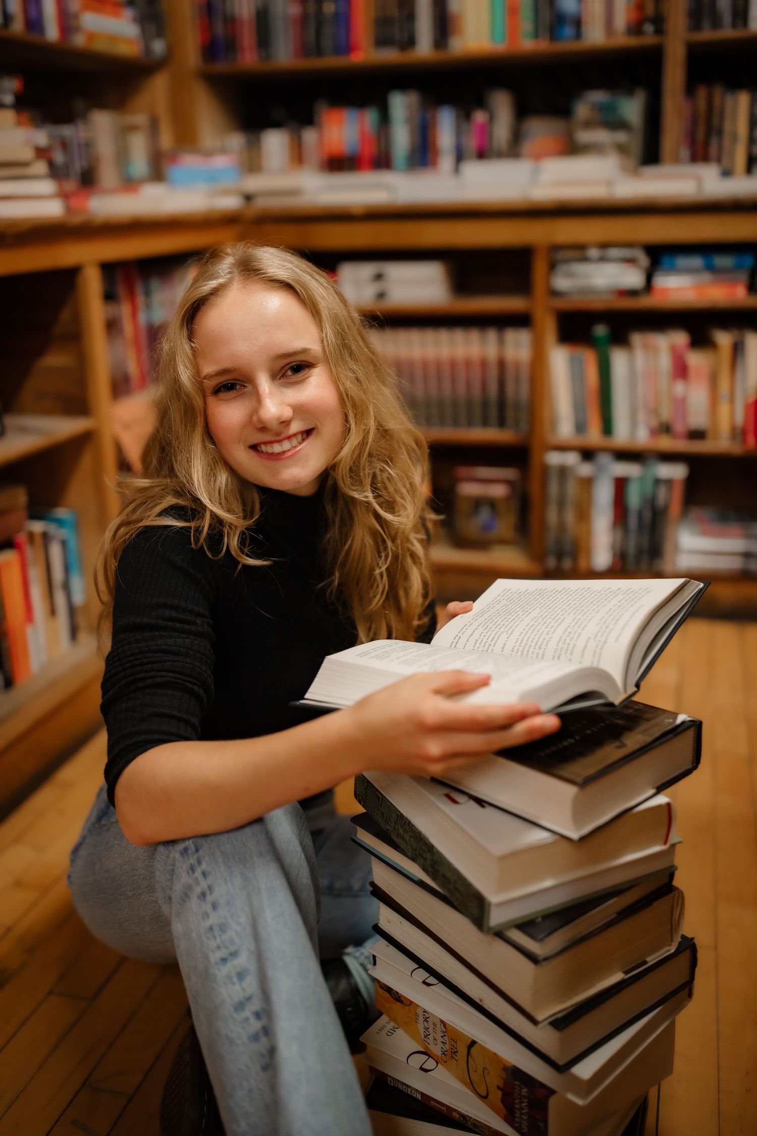 A reader sits comfortably beside a tall stack of books in a library setting.