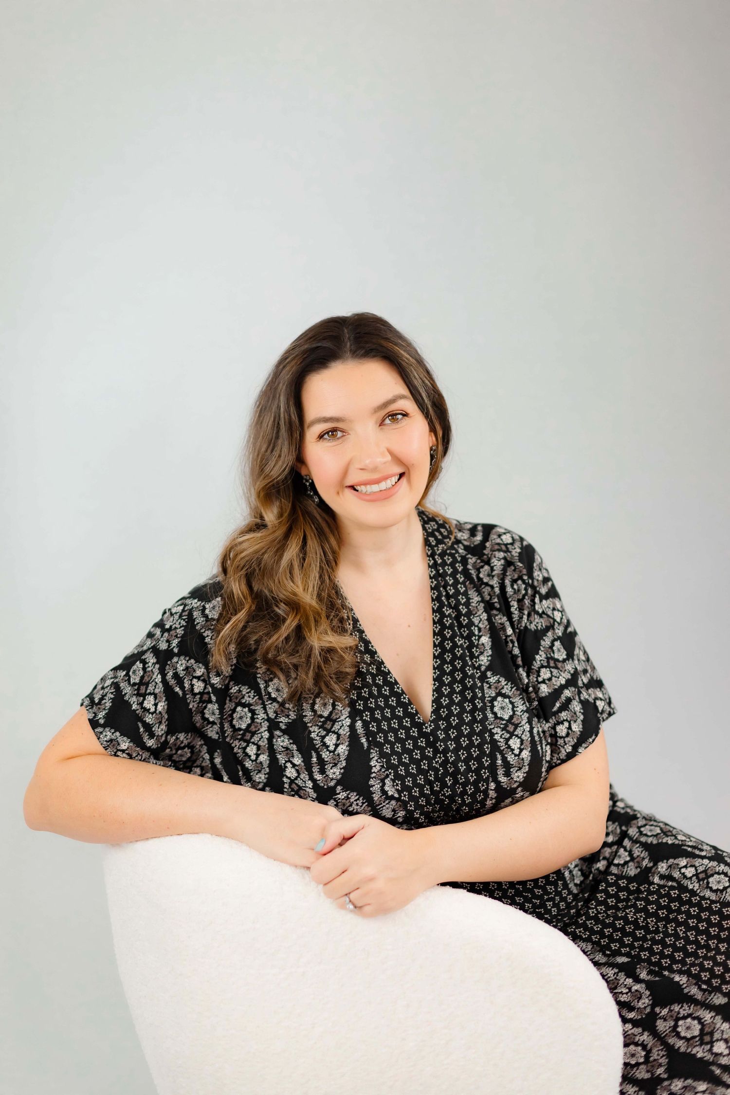 Portrait of woman in black patterned dress relaxing on white chair during photo session.