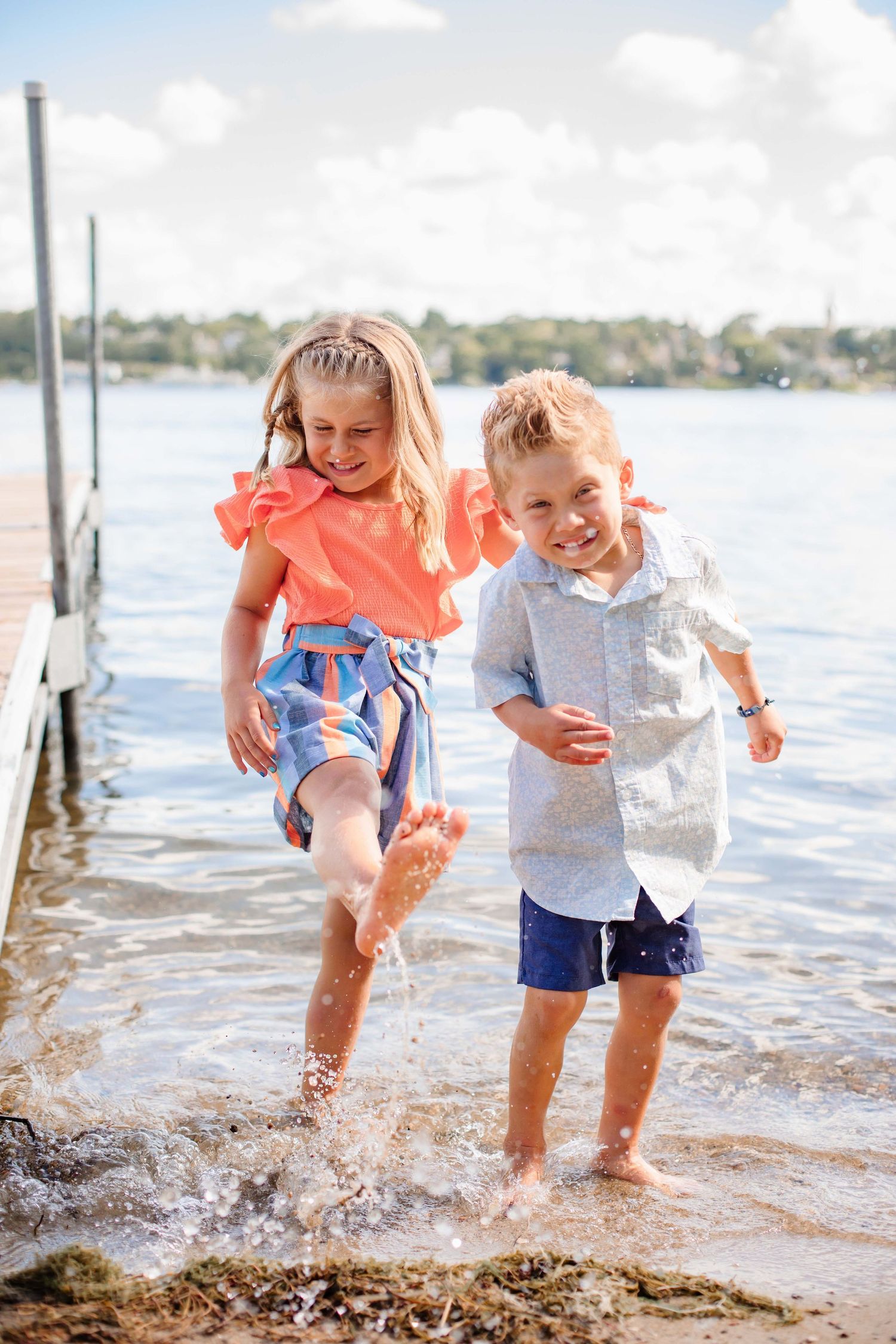 Children splashing and playing in lake water near wooden dock on sunny day.