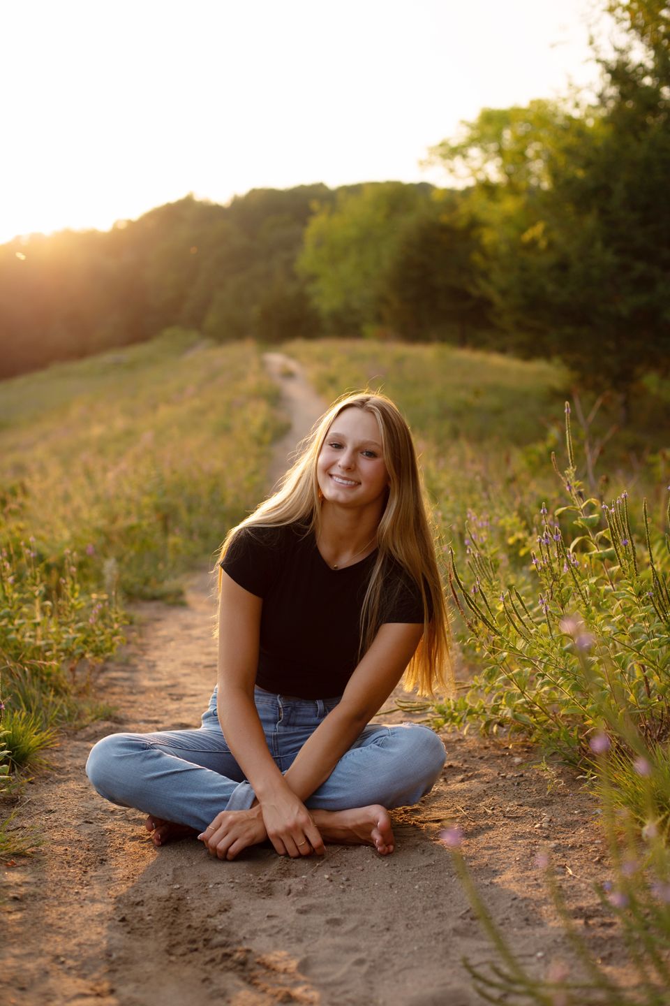 Person in casual attire sitting cross-legged on a dirt path during golden hour.