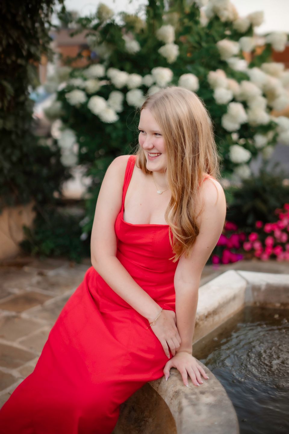 A woman in a red dress sits by a fountain surrounded by blooming white hydrangeas.