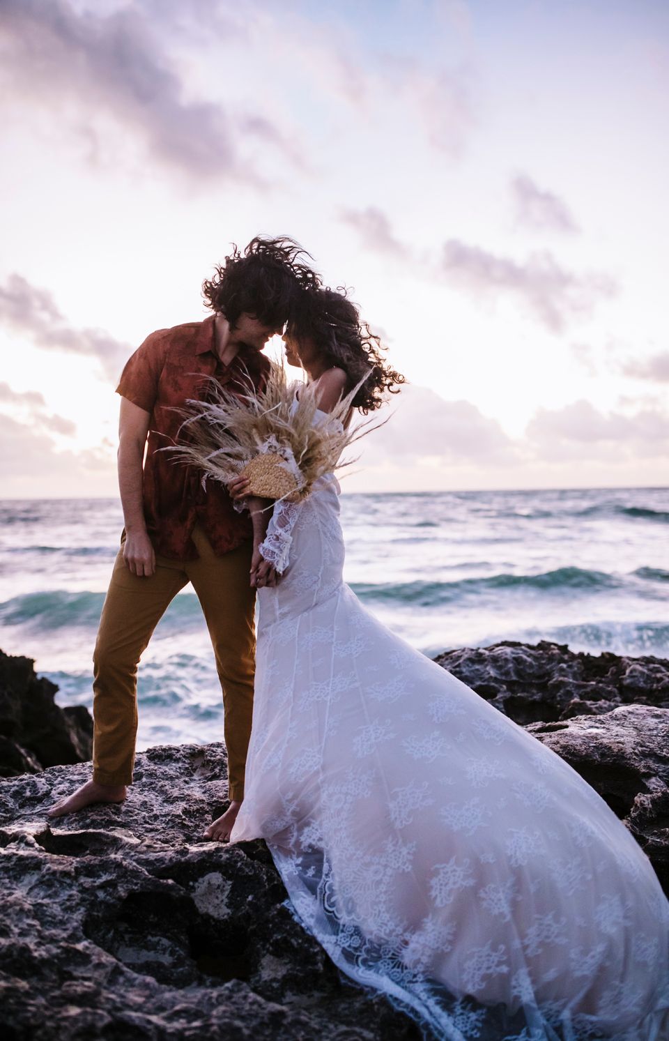 A couple in wedding attire shares a tender moment on ocean rocks at sunset with pampas grass bouquet.