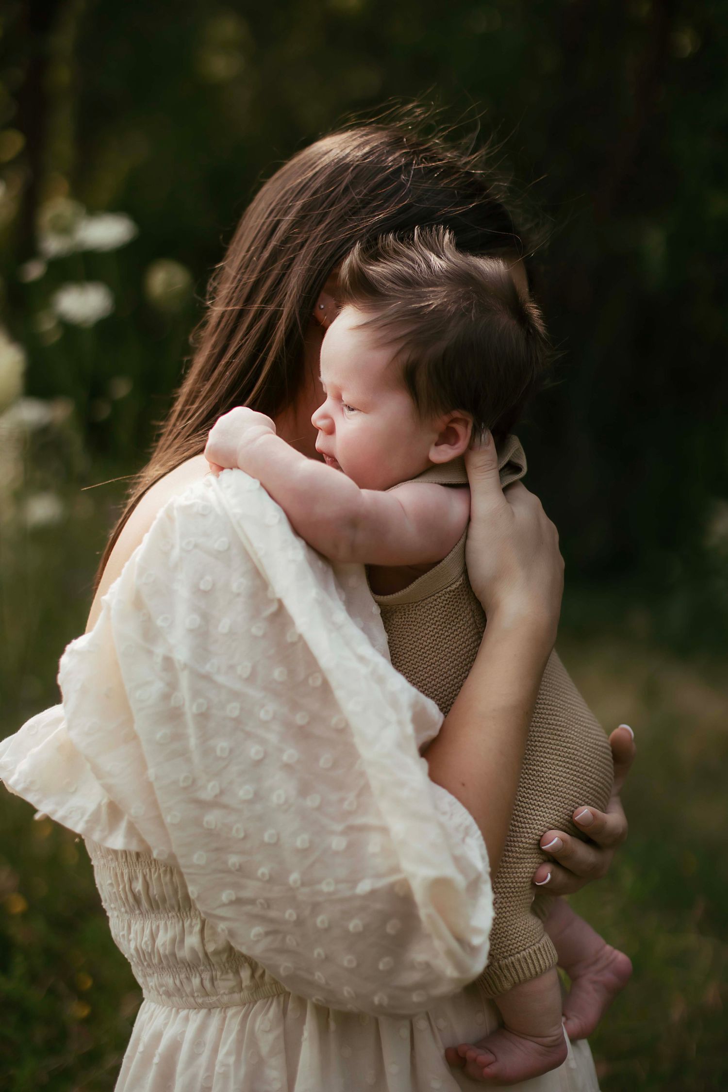 A mother tenderly holds her sleeping newborn baby outdoors, wearing a white dress in a lush green setting.
