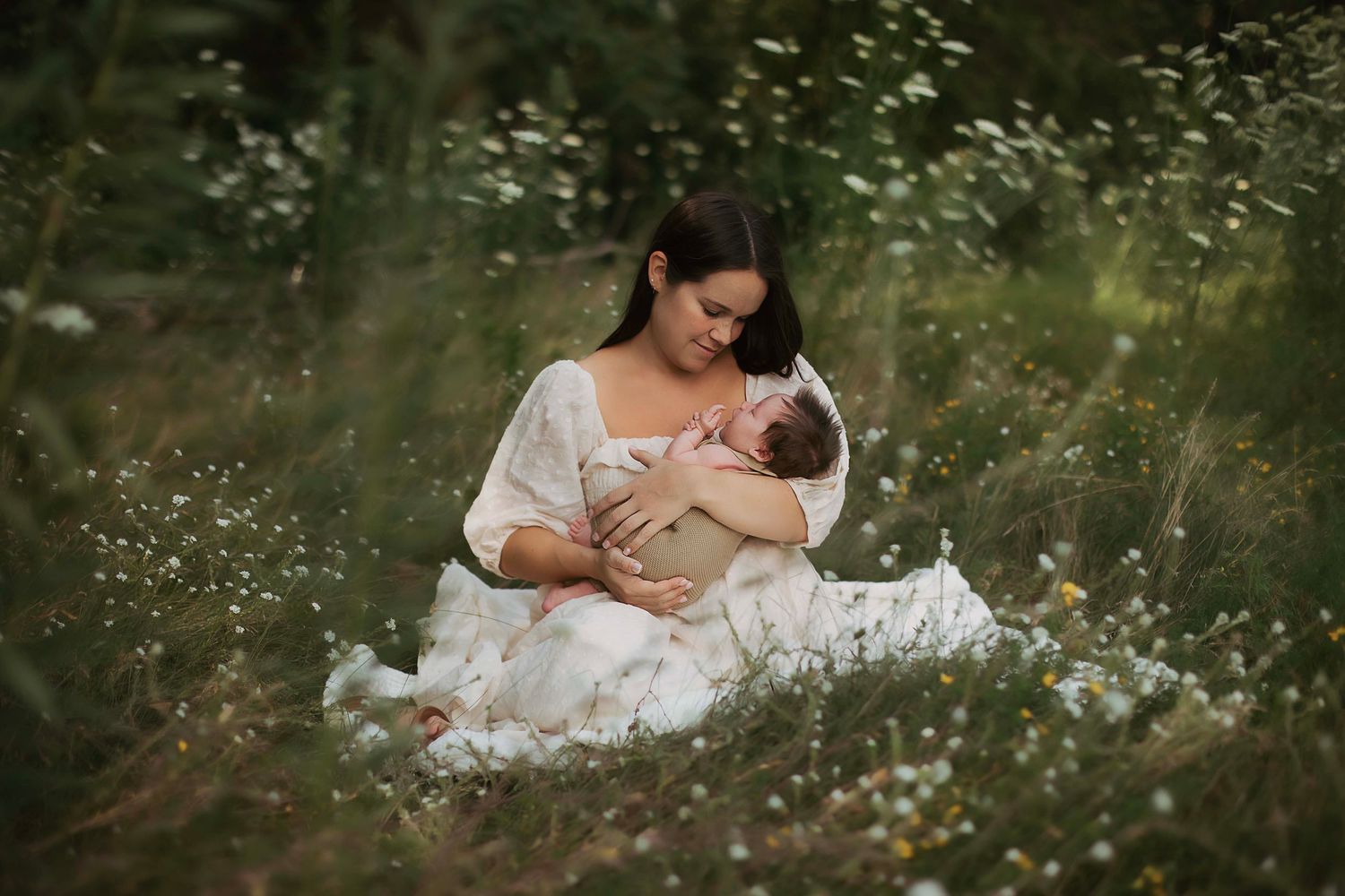 Mother in white dress sits in wildflower meadow cradling her newborn baby, surrounded by daisies and lush greenery.