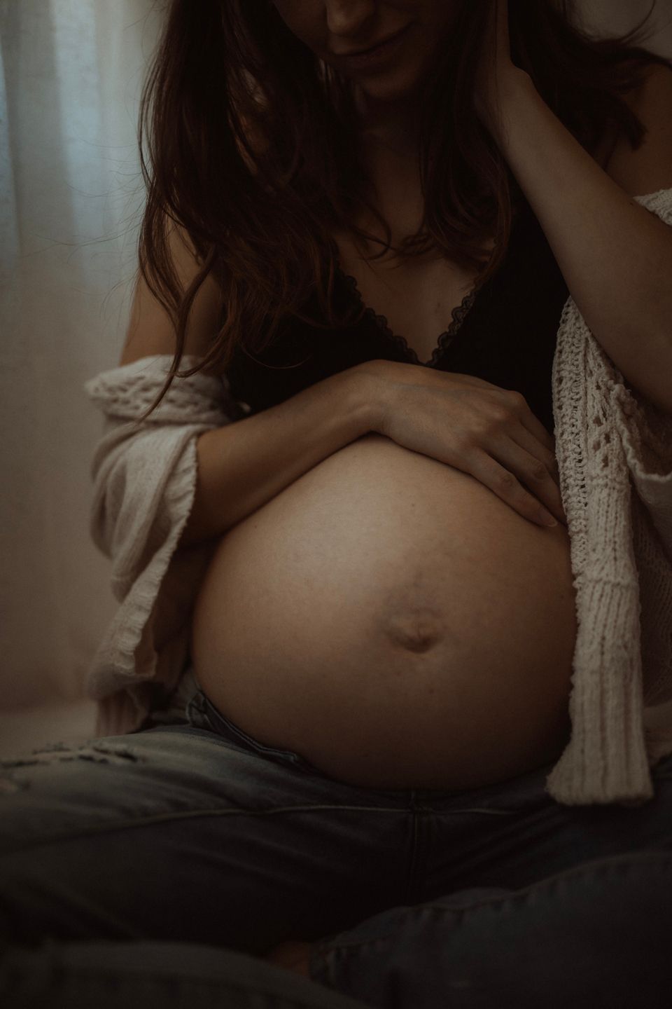 A moody maternity photo shows a pregnant belly with hands tenderly cradling it, wearing a black bra and white cardigan.