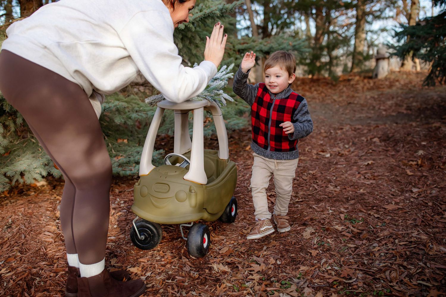 Adult decorating toy cozy coupe car with greenery while child watches in forest setting.