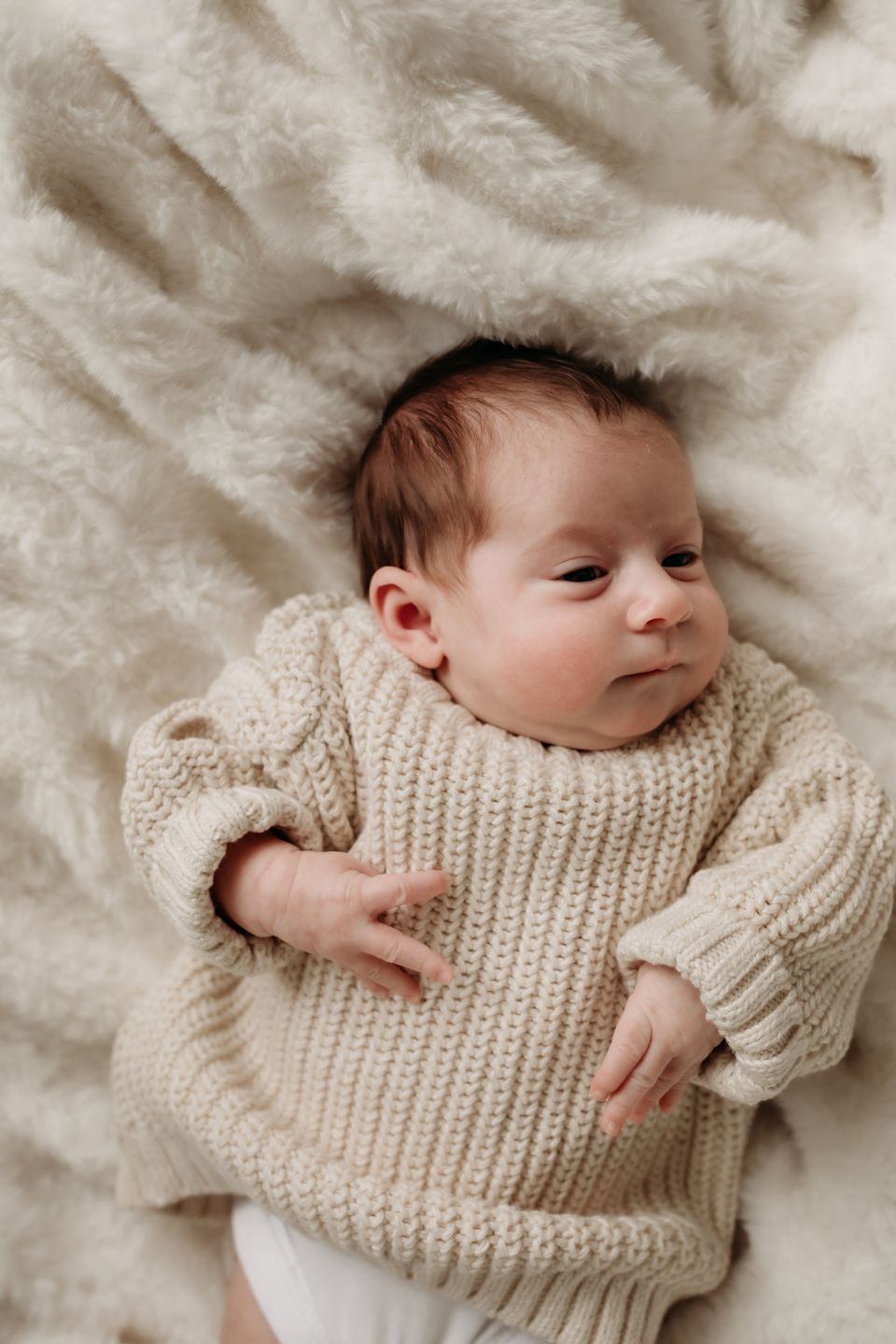 Newborn baby wearing cream knit sweater lying on plush white blanket.