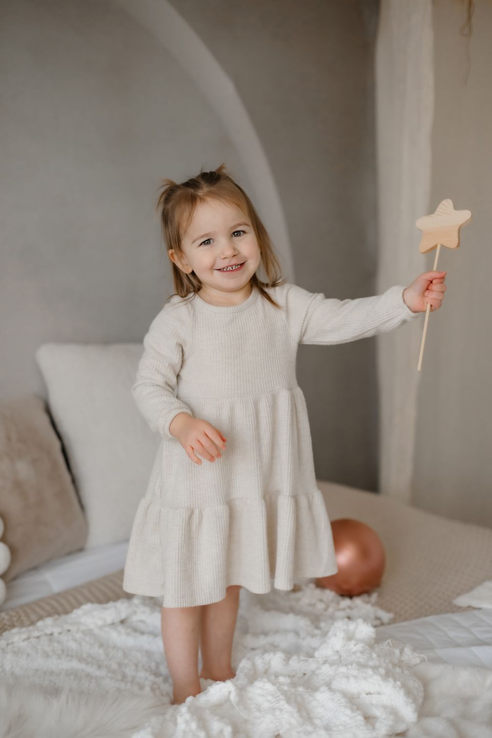 Child in a cream dress holds a star wand while standing on a white bed.