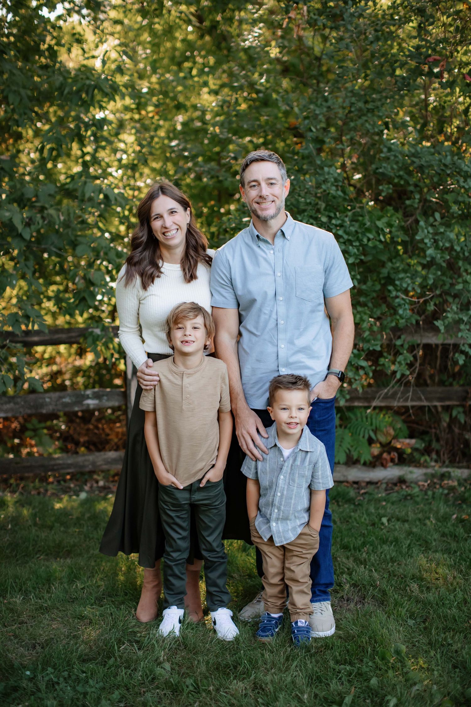 Family of four poses together for outdoor portrait against green foliage and wooden fence.