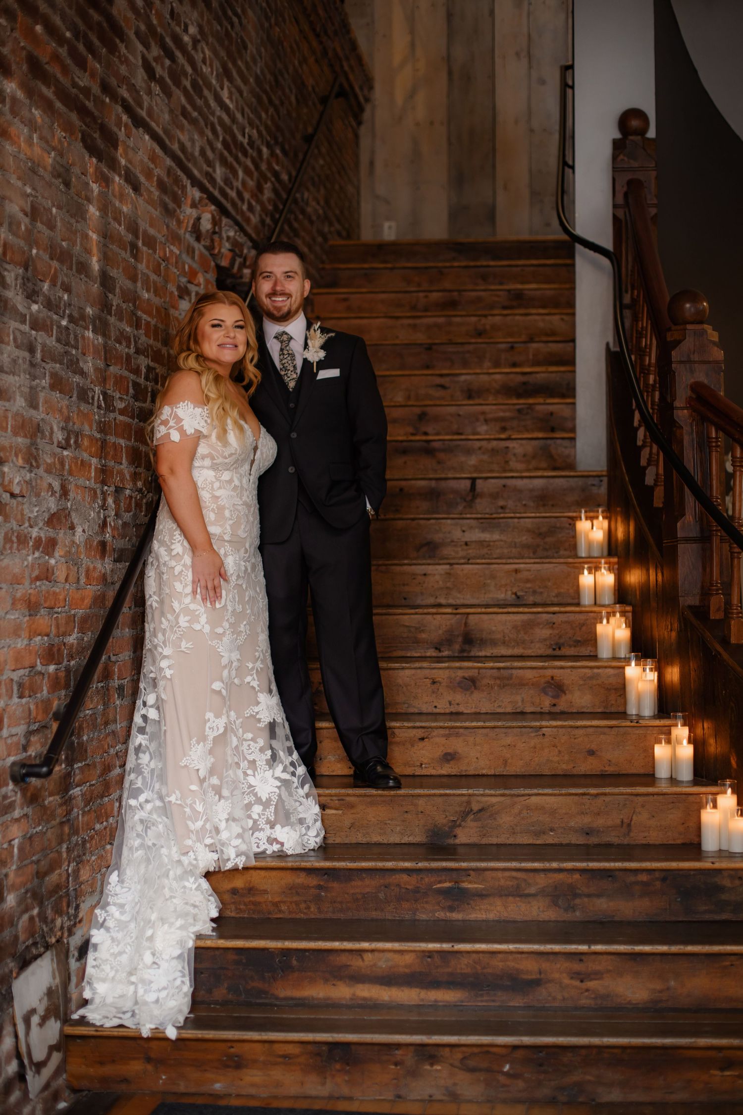 Wedding couple poses on rustic wooden staircase lit with romantic candles and exposed brick walls.