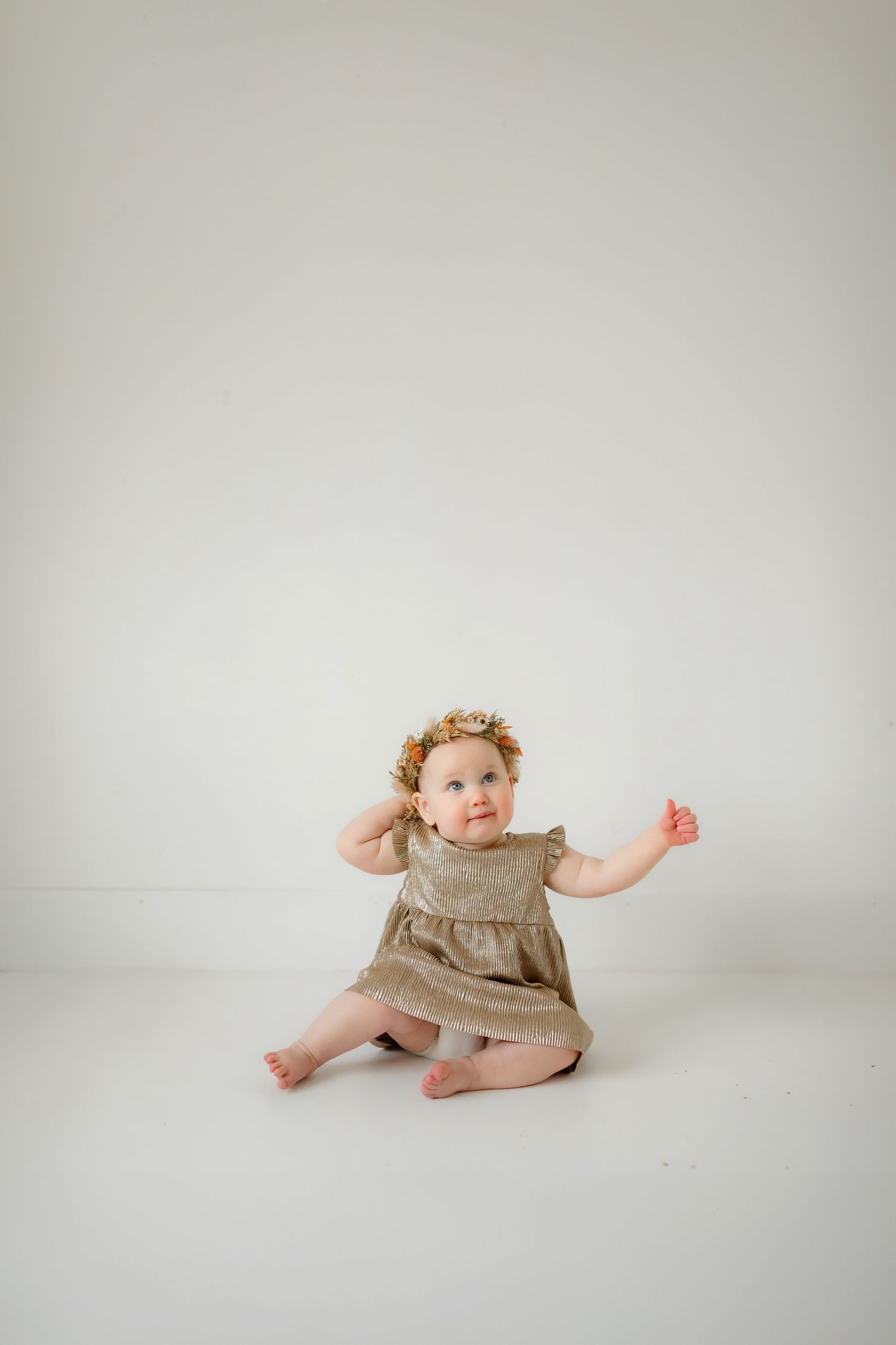 Baby in tan dress sits on white floor during minimalist portrait session.