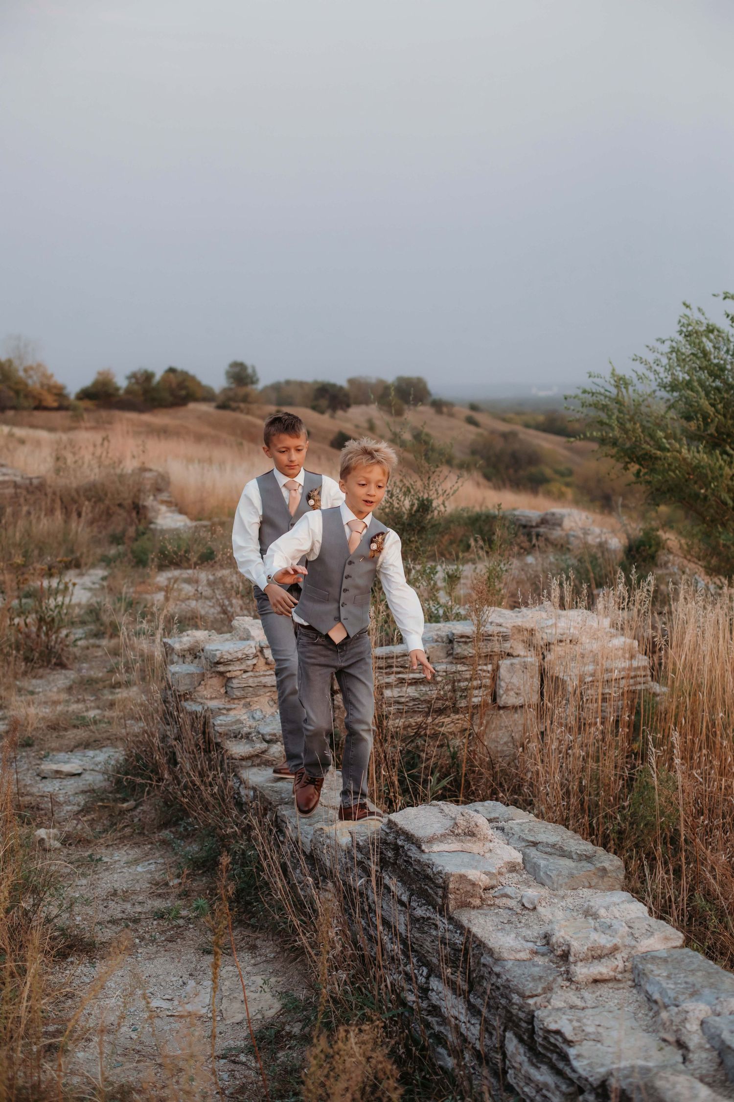 Young ring bearers in grey vests walking along stone wall in rustic outdoor setting.