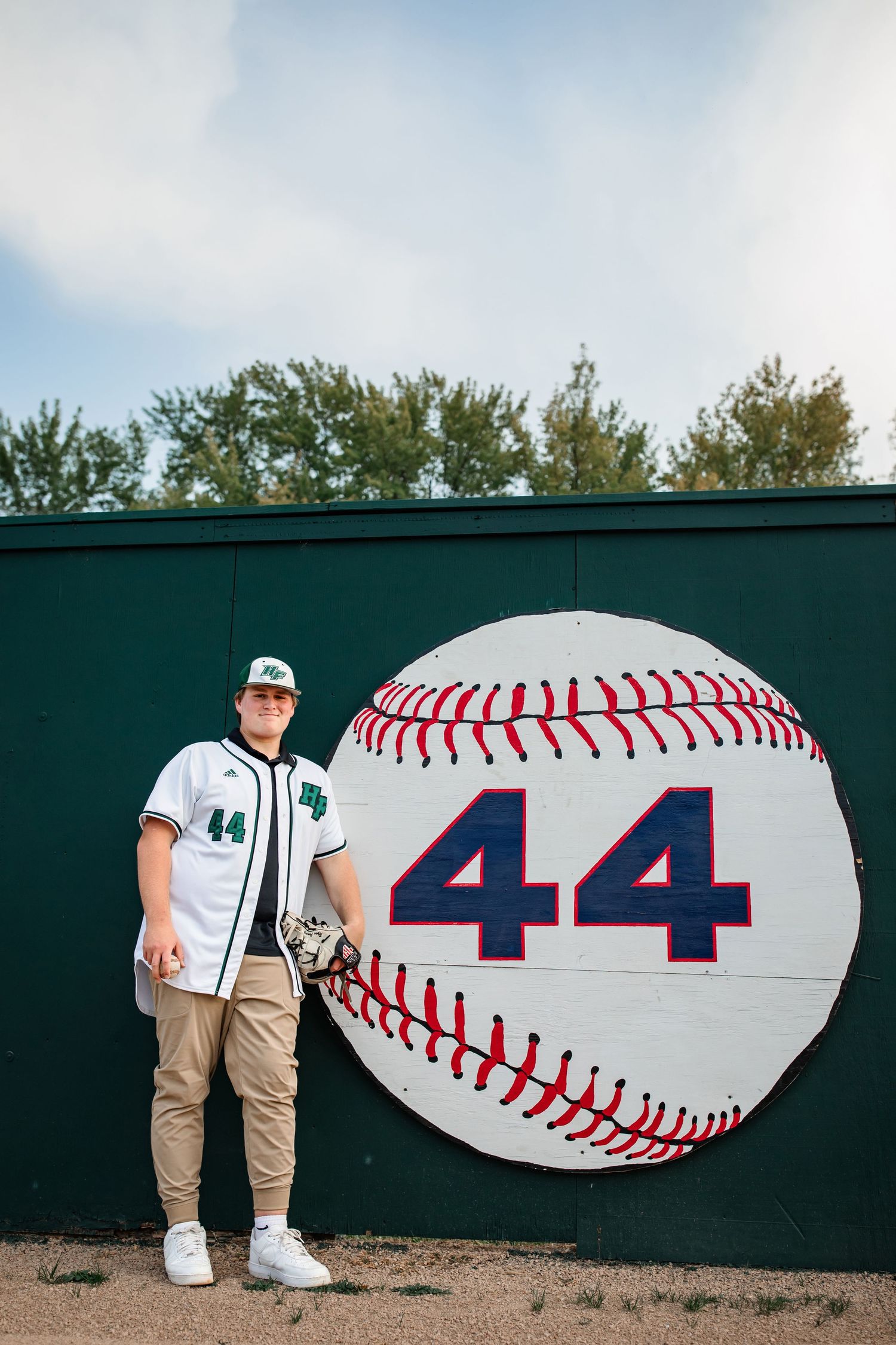Baseball player stands next to large baseball wall art displaying number 44 at sports field.