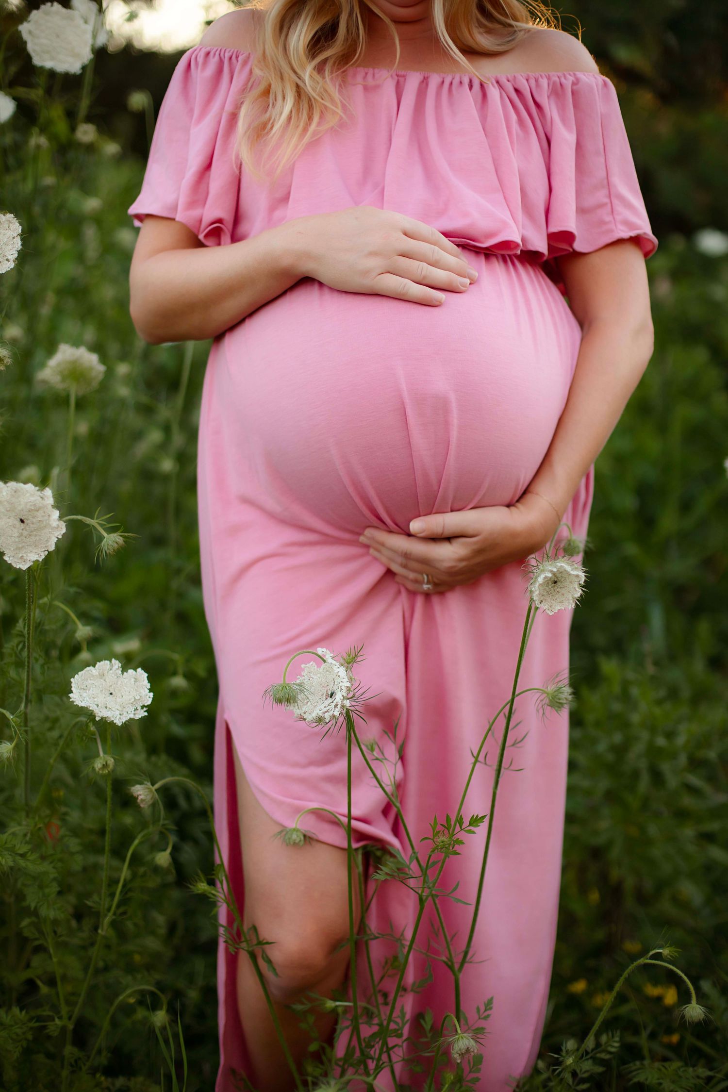 Pregnant woman in pink off-shoulder maxi dress cradling baby bump outdoors among white wildflowers.