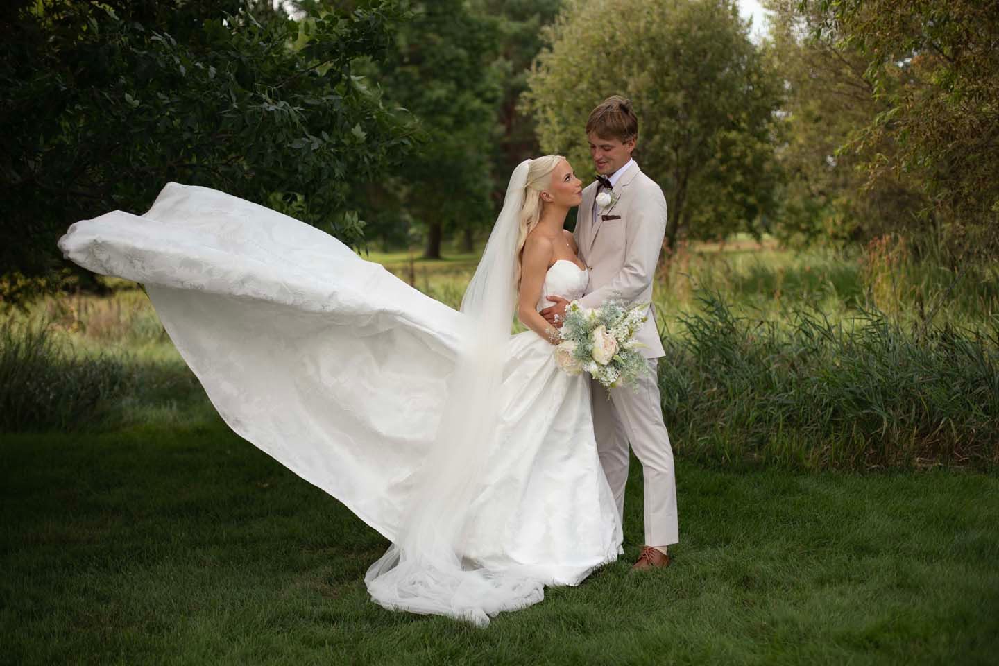 Bride in flowing white gown with veil and groom in tan suit embrace outdoors on green lawn with trees.
