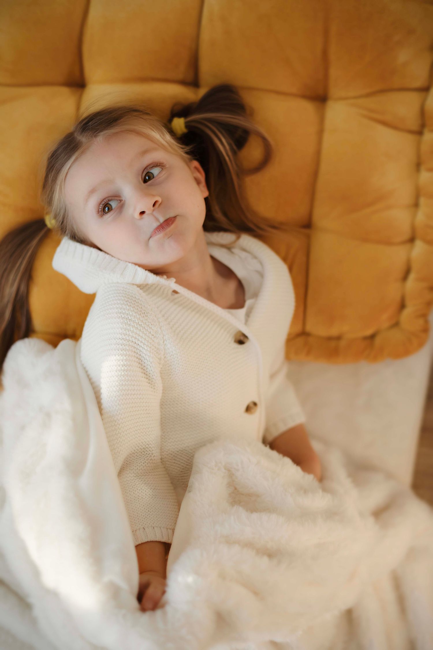 A child wearing a white knit sweater lies on a beige couch while making playful expressions.