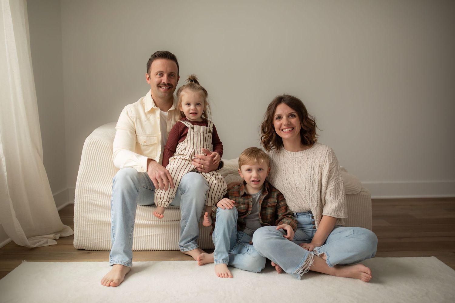 Family relaxes together on floor against neutral wall in coordinated casual outfits.