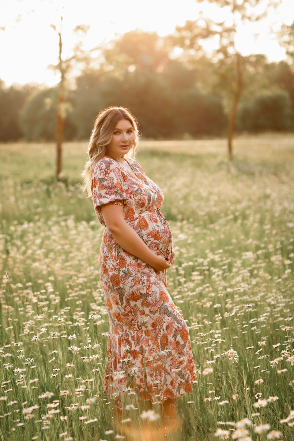 An expectant woman in a floral dress poses amid a meadow of daisies at sunset.