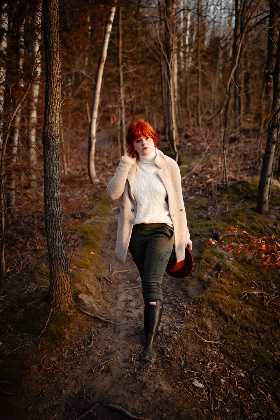 A person walks along a forest path wearing a beige coat and rain boots in autumn.