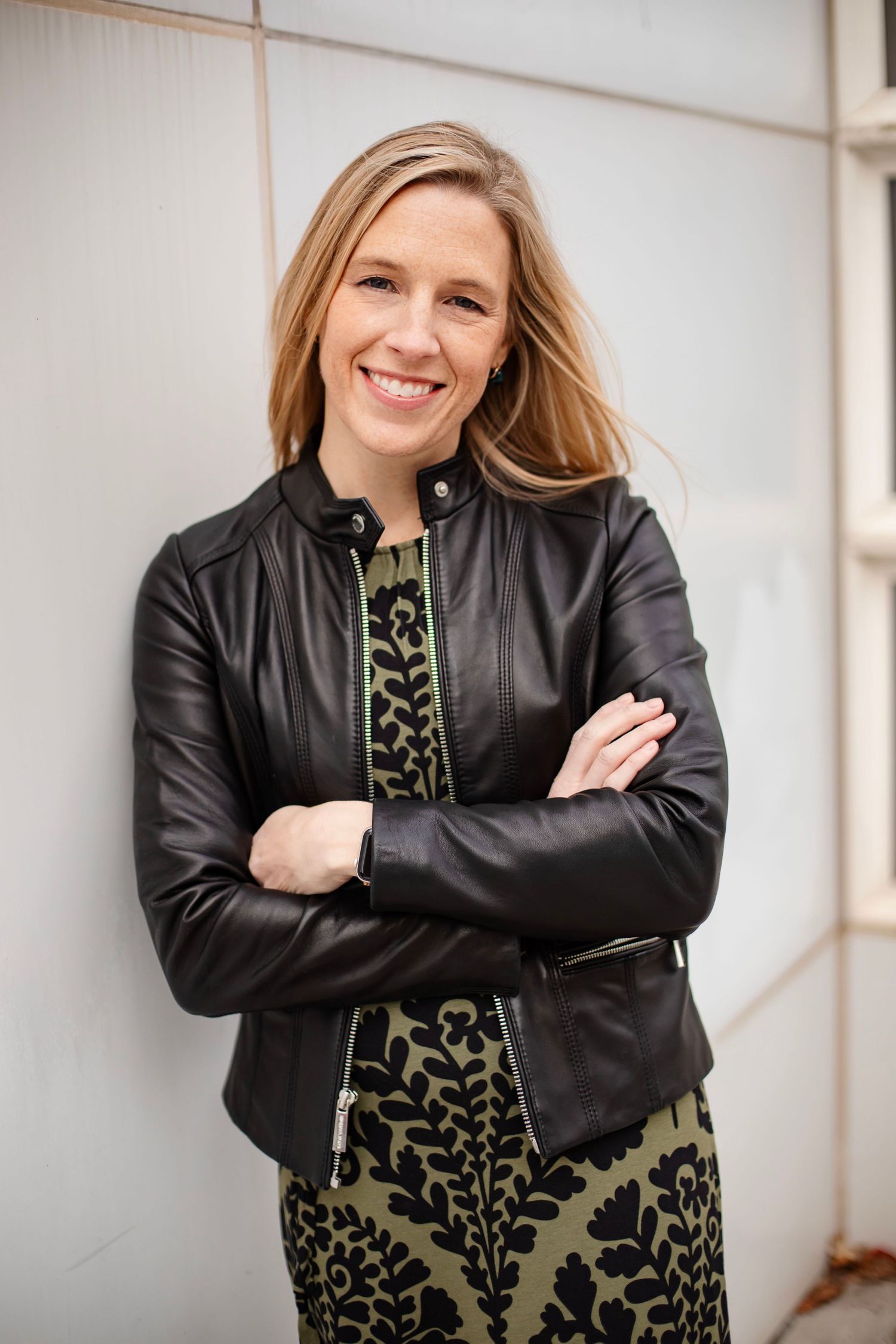 Professional portrait featuring black leather jacket and patterned dress against white wall.