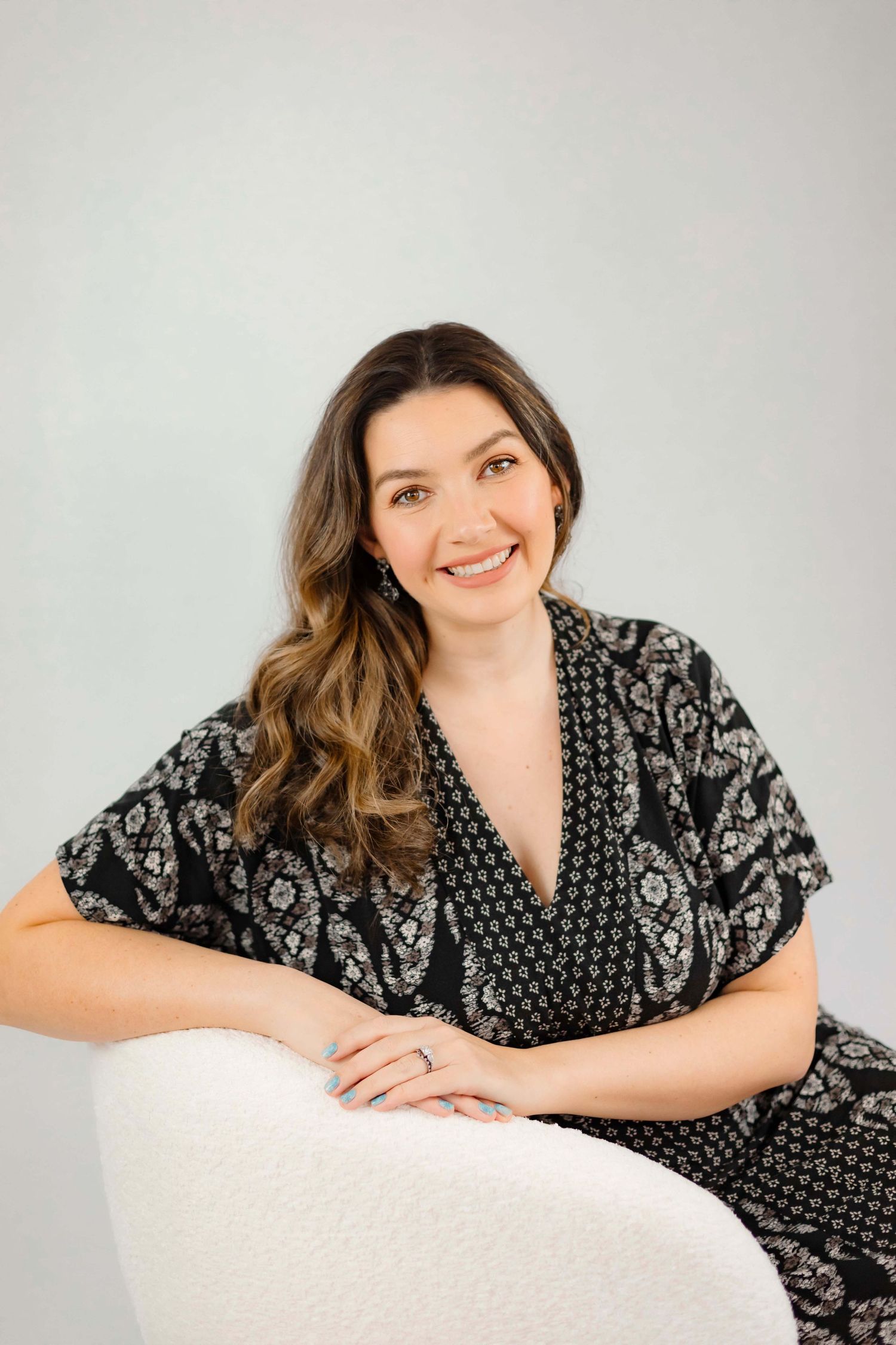 Woman in black floral dress sits comfortably on white chair during studio photoshoot.