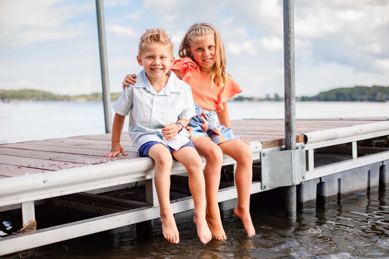 Two children sit swinging their legs on dock edge over lake water.