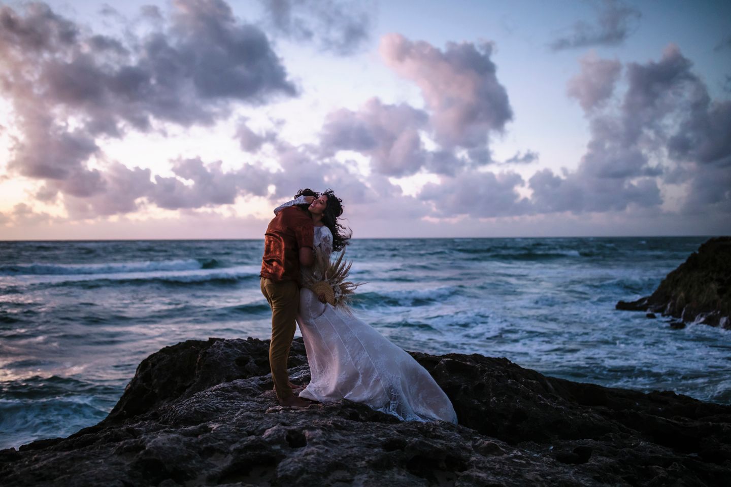 Silhouetted couple embraces on rocky coastline during sunset with dramatic waves crashing behind them.
