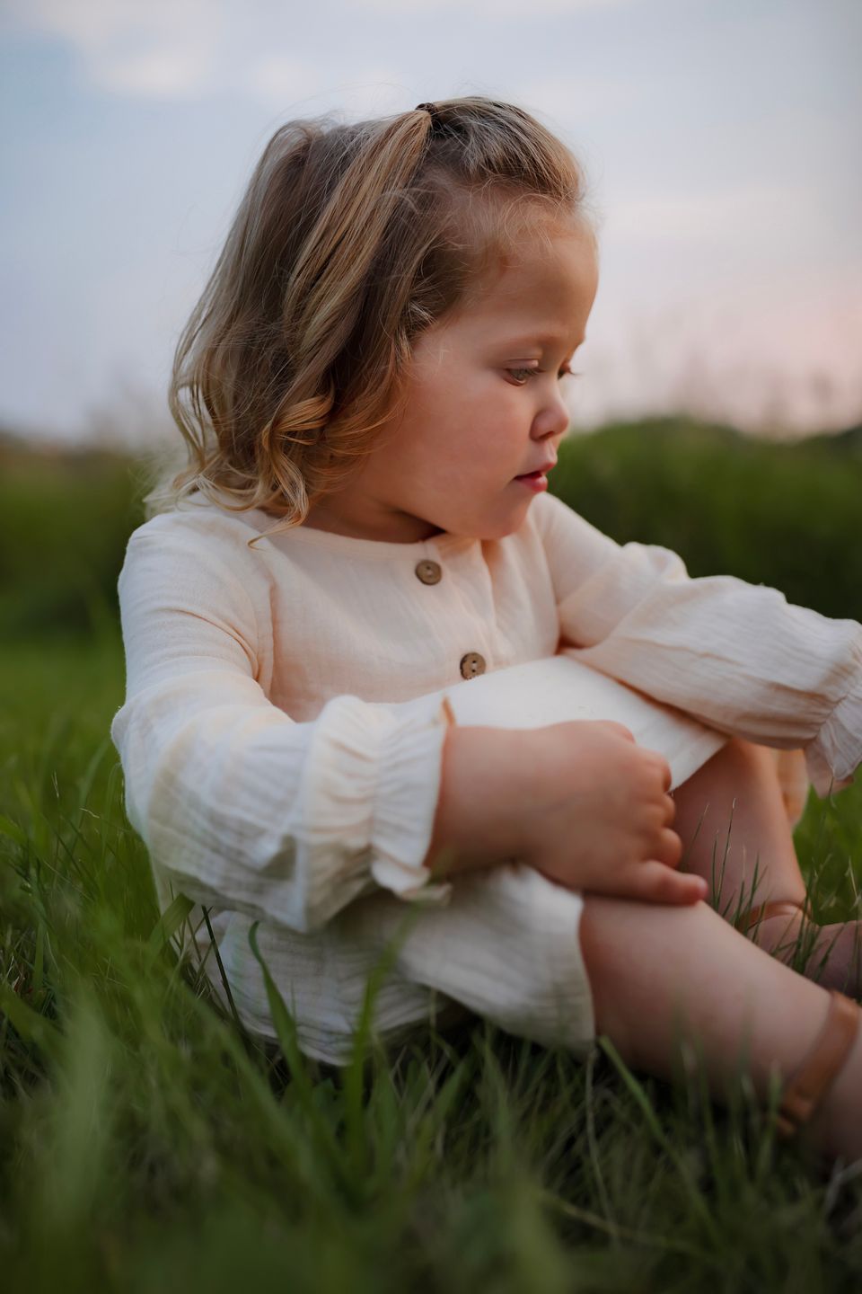 A person in a cream button-up dress sits thoughtfully in tall grass.
