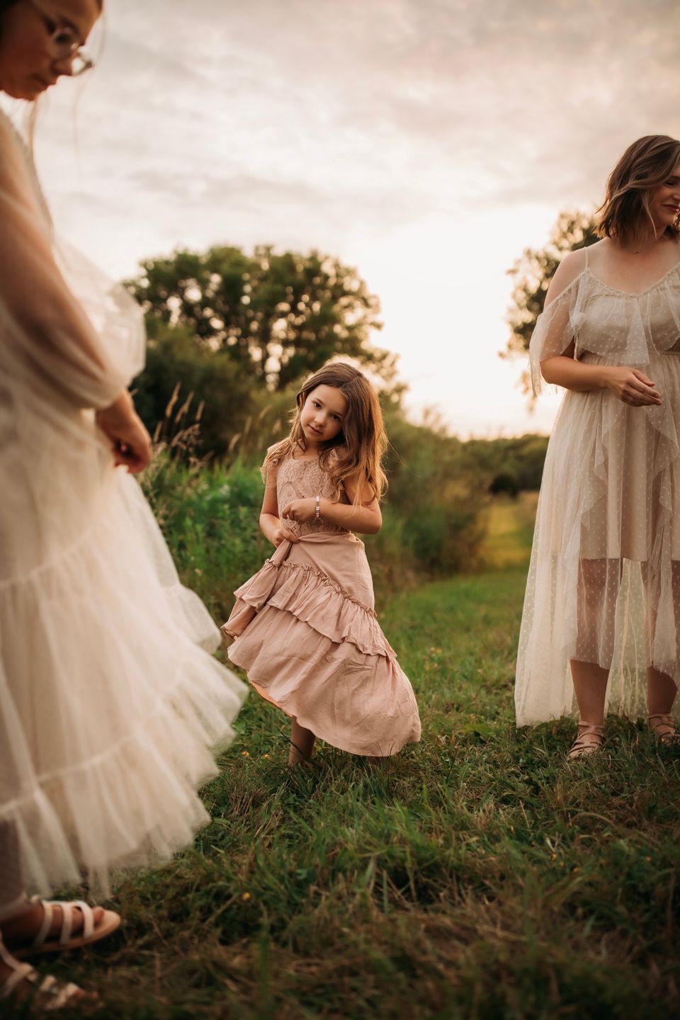 Group in flowing dresses twirls and dances together in a summer field at dusk.