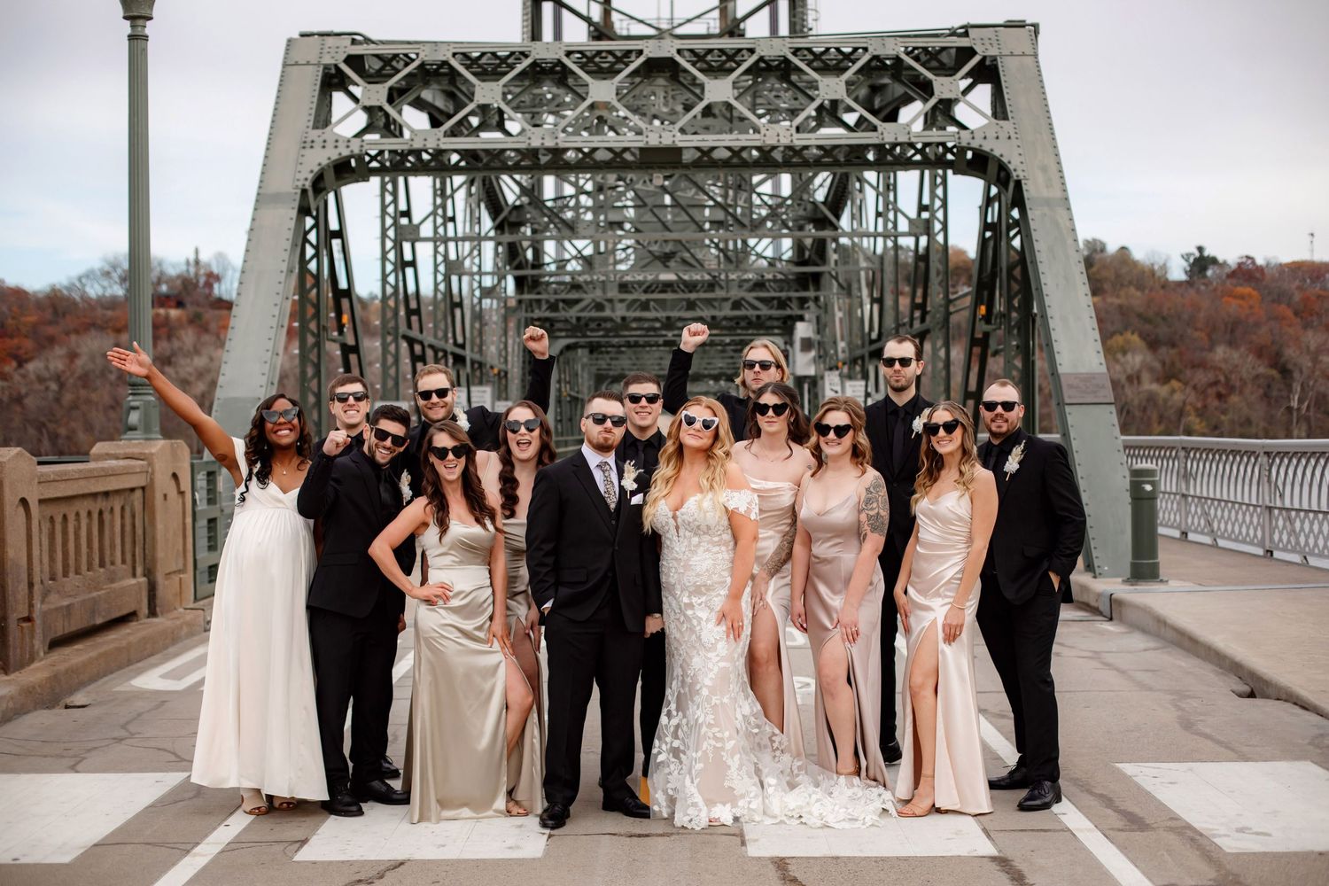 Full wedding party in formal attire poses on historic steel bridge structure.