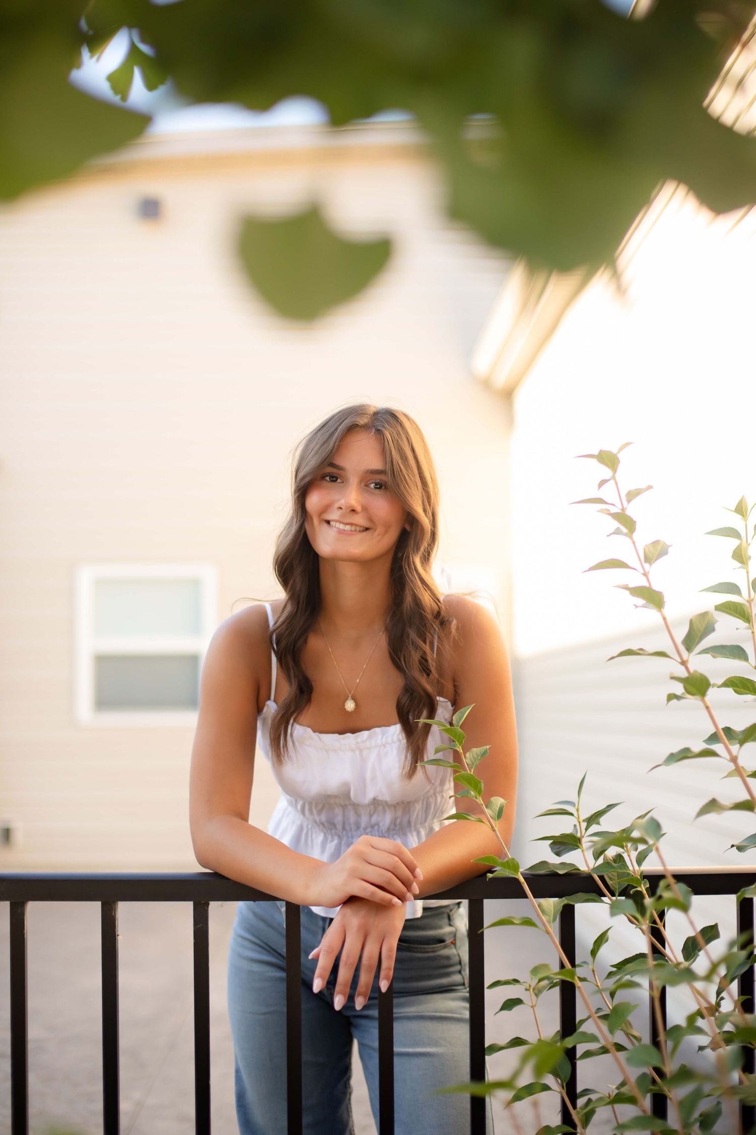 Young woman in white top and jeans leaning on black railing outdoors during golden hour.