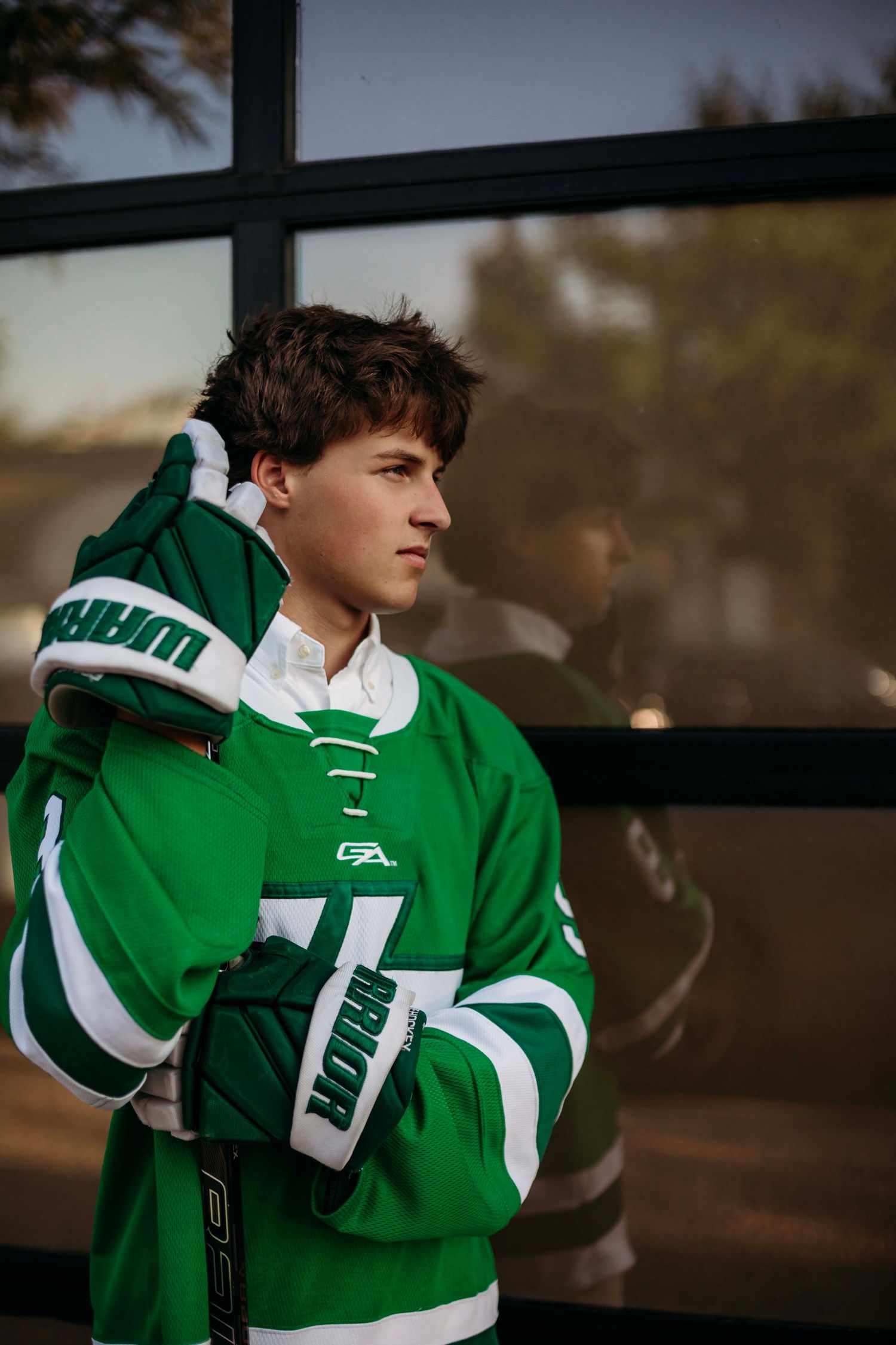 Young hockey player in green Warrior jersey holding gloves near glass window.