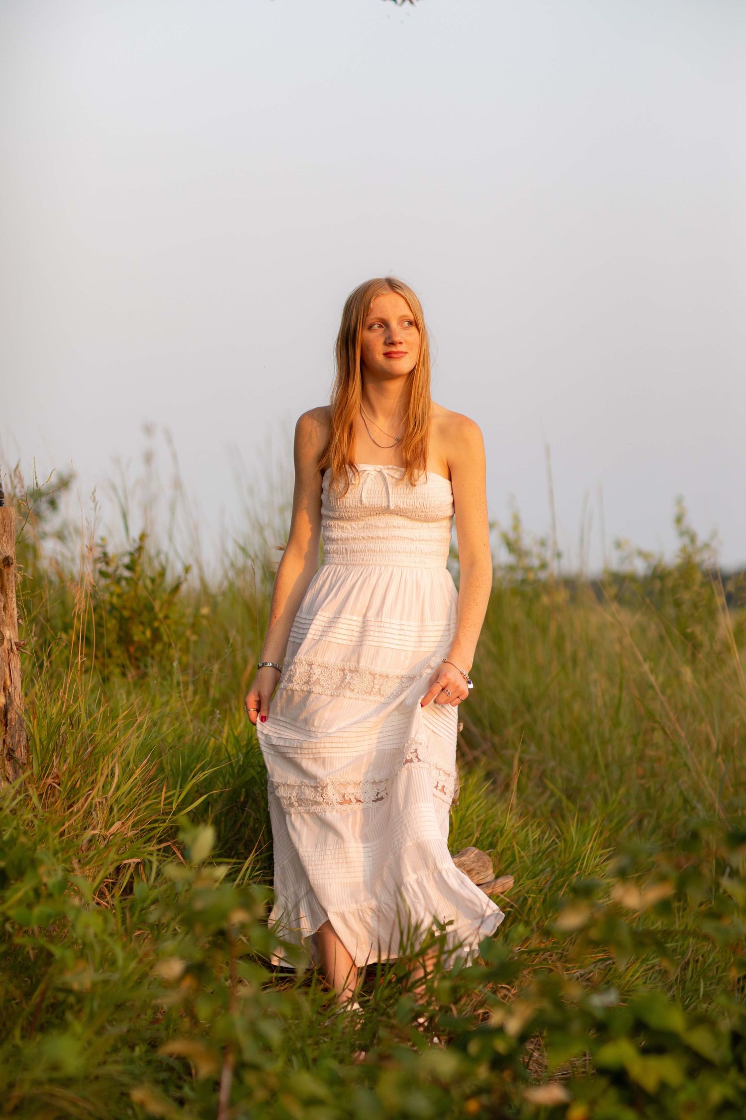 Young woman in white sundress standing in green field during golden hour sunset light.