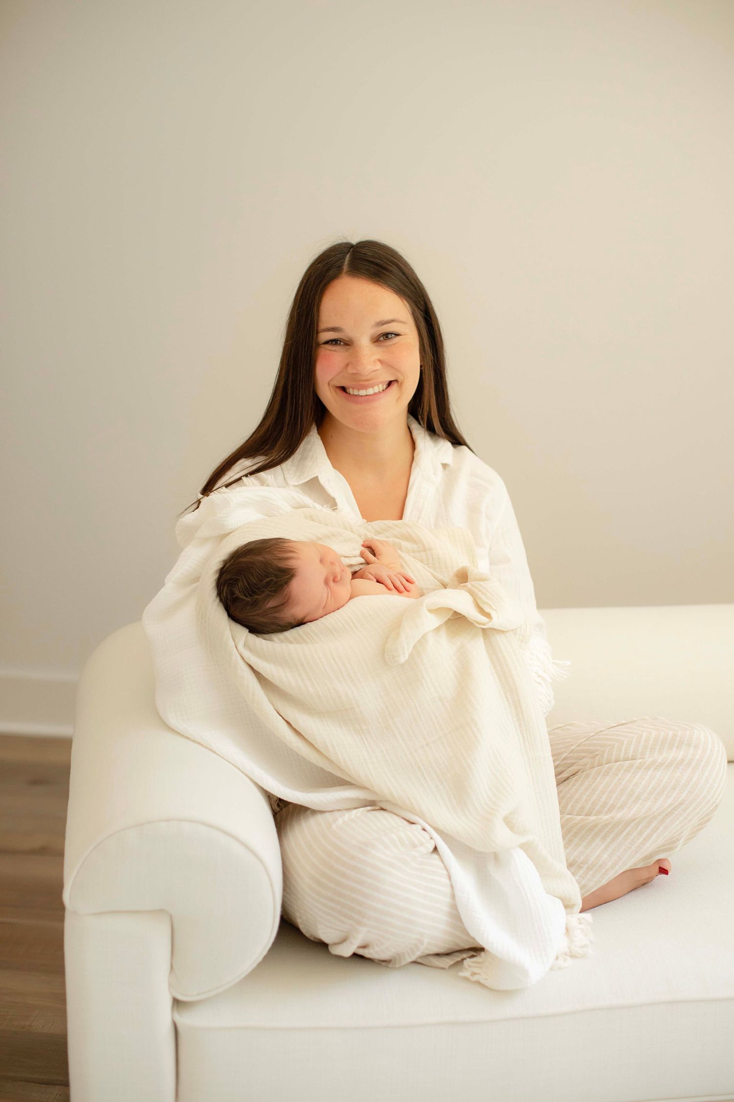 Smiling mother sitting on white sofa holding sleeping newborn wrapped in cream blanket.