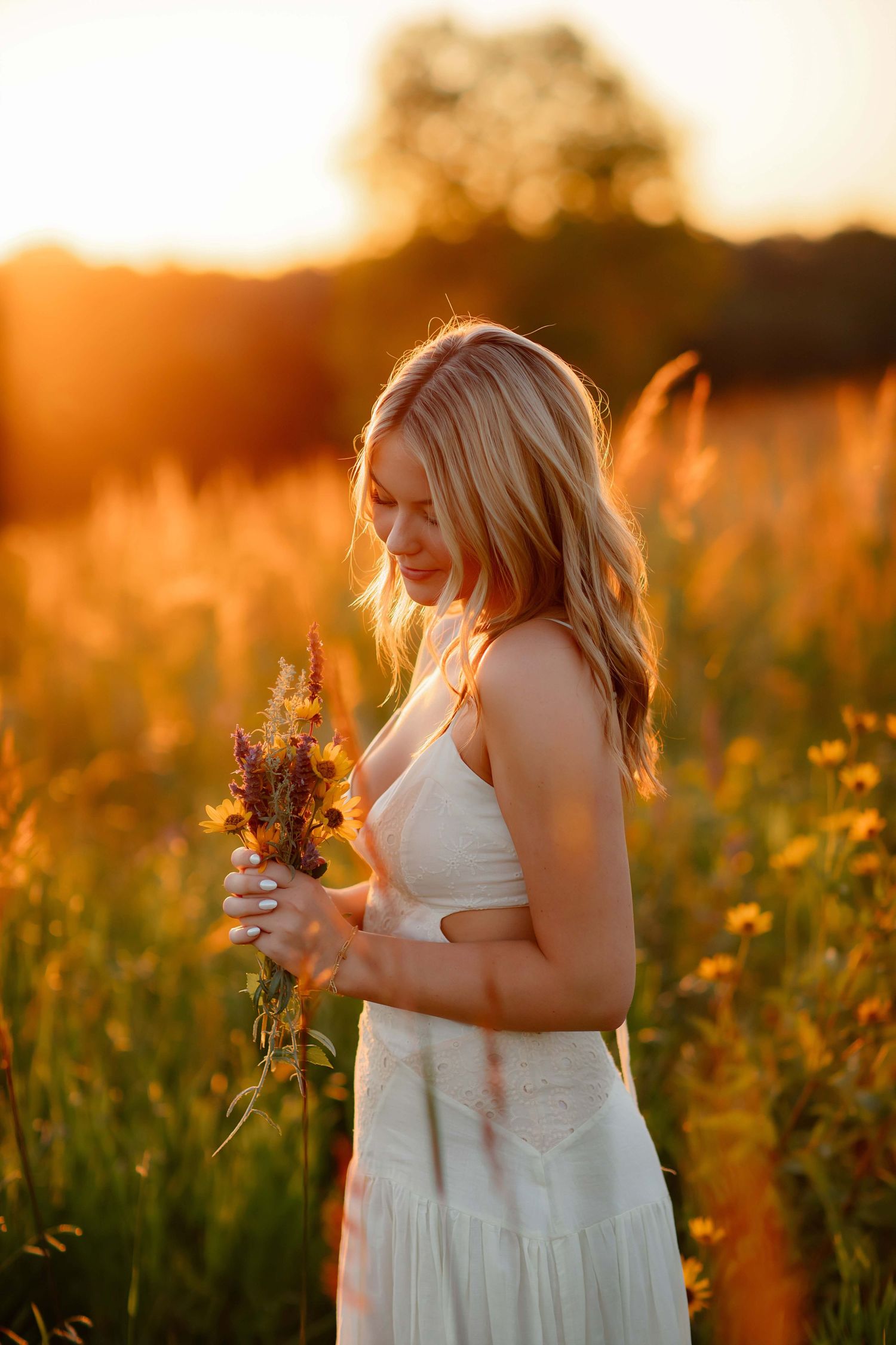 A high school senior in a white dress stands in a golden wildflower field at sunset holding dried flowers.