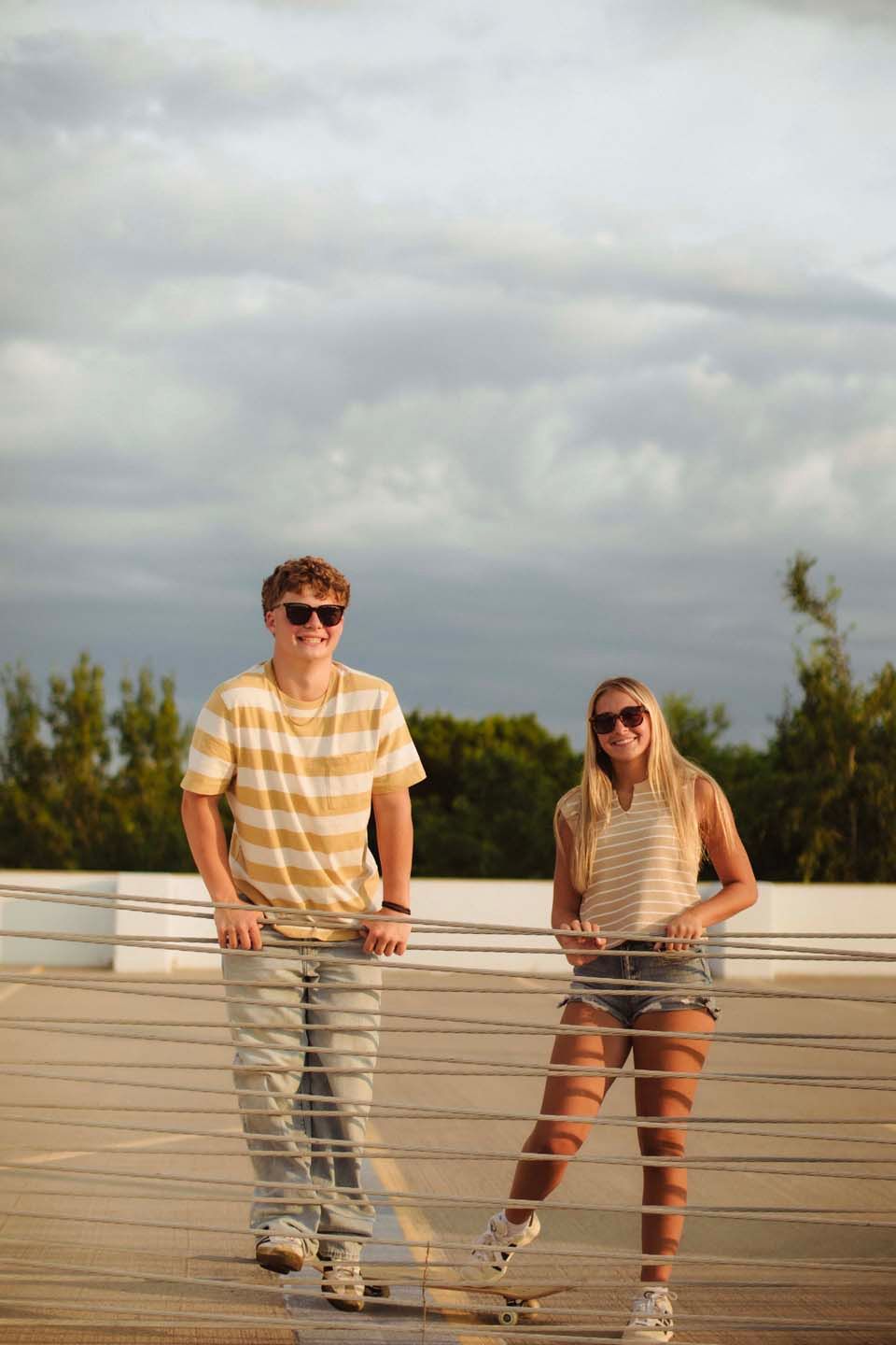 Two teens in matching striped shirts and sunglasses pose with skateboards on a bridge at sunset.