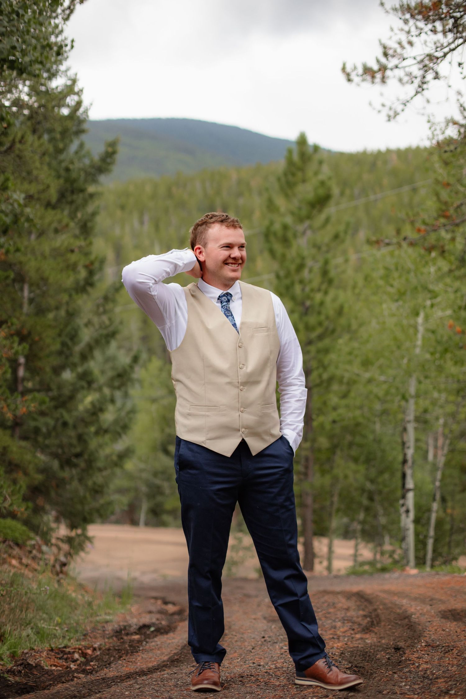 Groom in tan vest and navy pants poses playfully on a dirt path surrounded by evergreen trees in mountain setting.