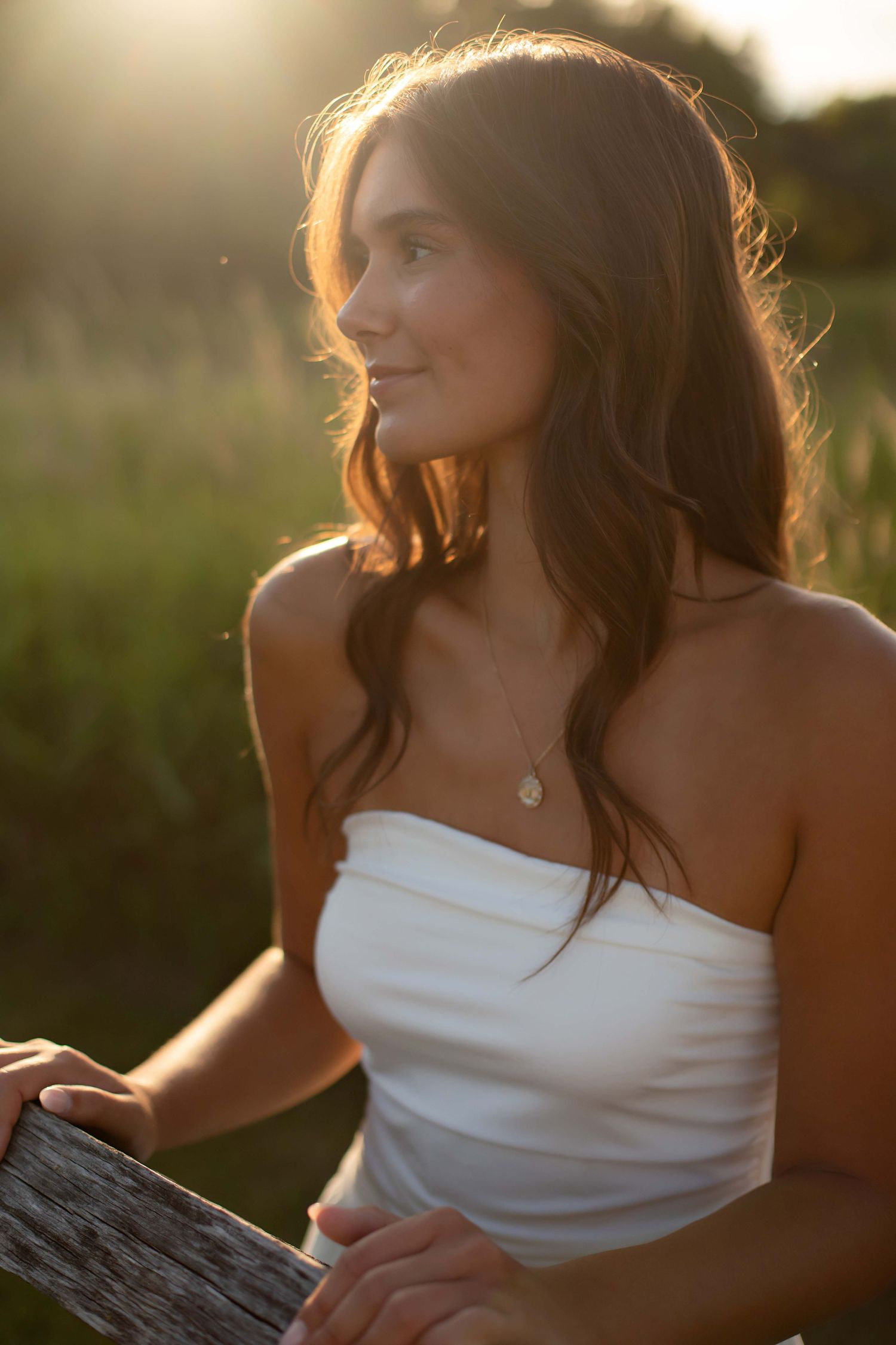 Young woman in a white strapless dress bathed in golden sunset light, standing by a rustic wooden fence.